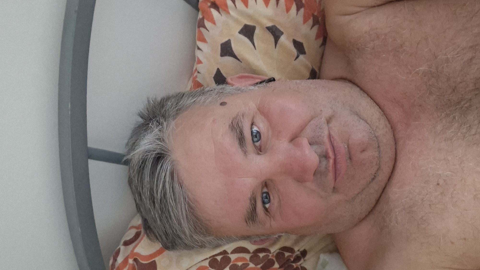 A close-up, overhead shot shows a fair-skinned, middle-aged man with graying hair and blue eyes. He is lying down, with his head tilted slightly to the right. His skin is fair and shows some signs of aging. He has a mole on his left cheek. He is wearing a pillow with an abstract pattern of orange and brown shapes. The background is a plain white wall. The man's chest and neck are visible, and he appears to be shirtless. The lighting is soft and diffused, suggesting an indoor setting, possibly a bedroom. There is no indication of the city of Hîncești, Moldova, or any other specific location in the image.