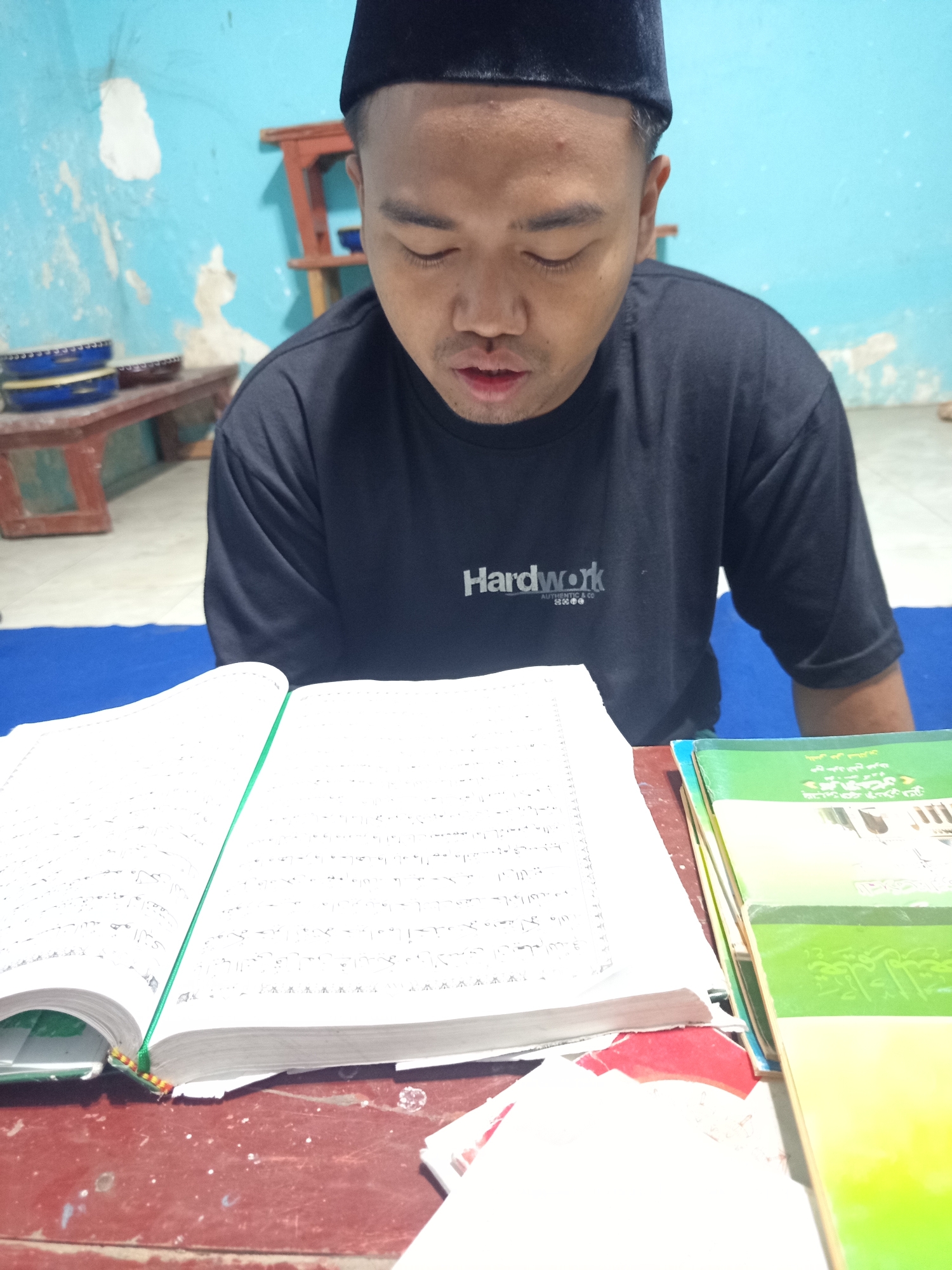 A man wearing a black peci and a black t-shirt with "Hardwork" printed on it is sitting at a red wooden table, looking down at an open book. The book appears to be a religious text with handwritten notes. To the right of the open book are several other books stacked on top of each other, with green and yellow covers. The background is a light blue wall with patches of white paint. In the background on the left, there are two tambourines on a wooden bench. The scene is indoors, likely in a home or place of worship, and based on the provided context, this location is the city of Ngablak, Indonesia.