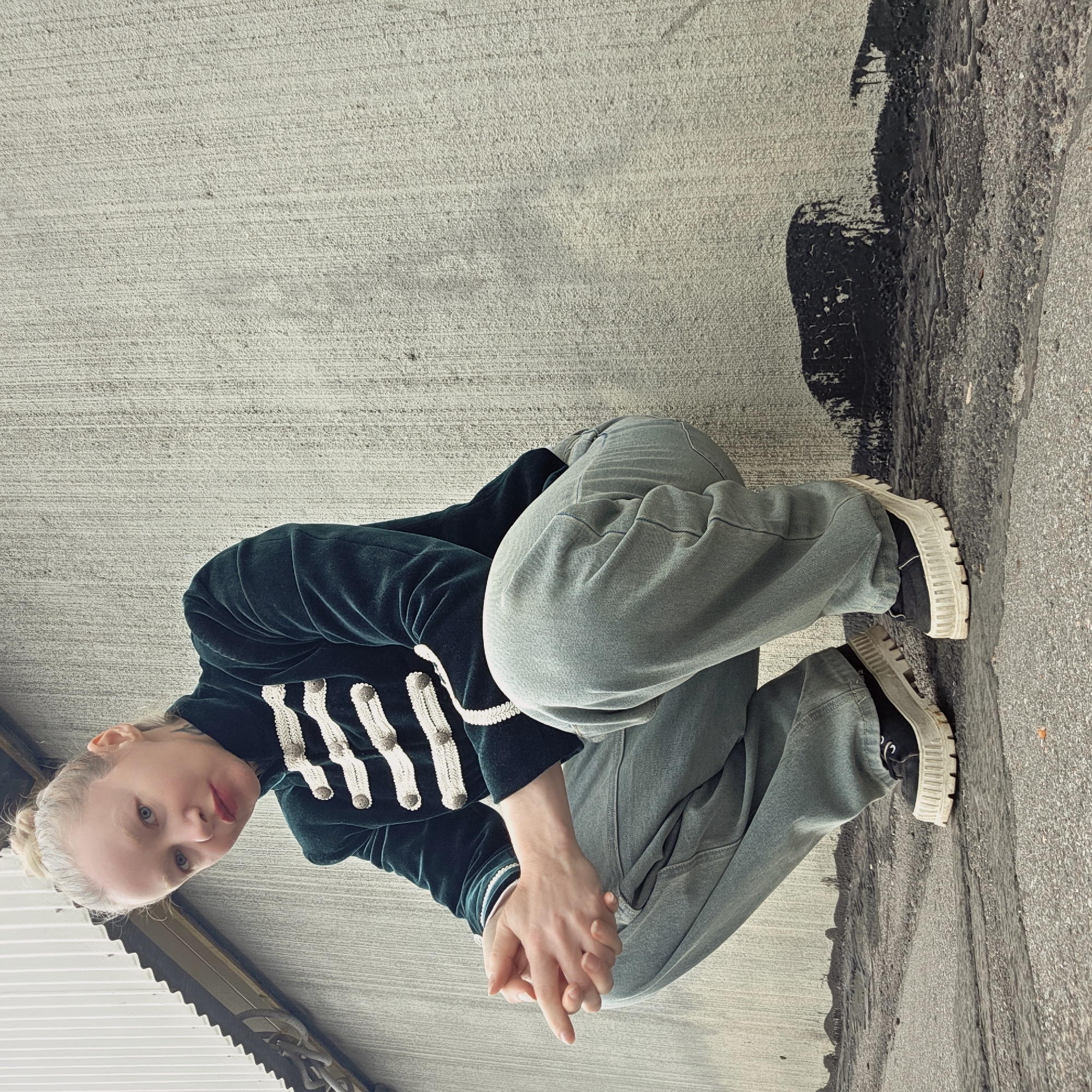 A fair-skinned person with pale blonde hair pulled back is crouching on a concrete surface. They are wearing light green jeans and a dark green velvet jacket adorned with white embellishments down the front. Their hands are clasped together in front of them. The concrete surface is stained with a broad black streak. The location may be in Saint Petersburg, Russia.
