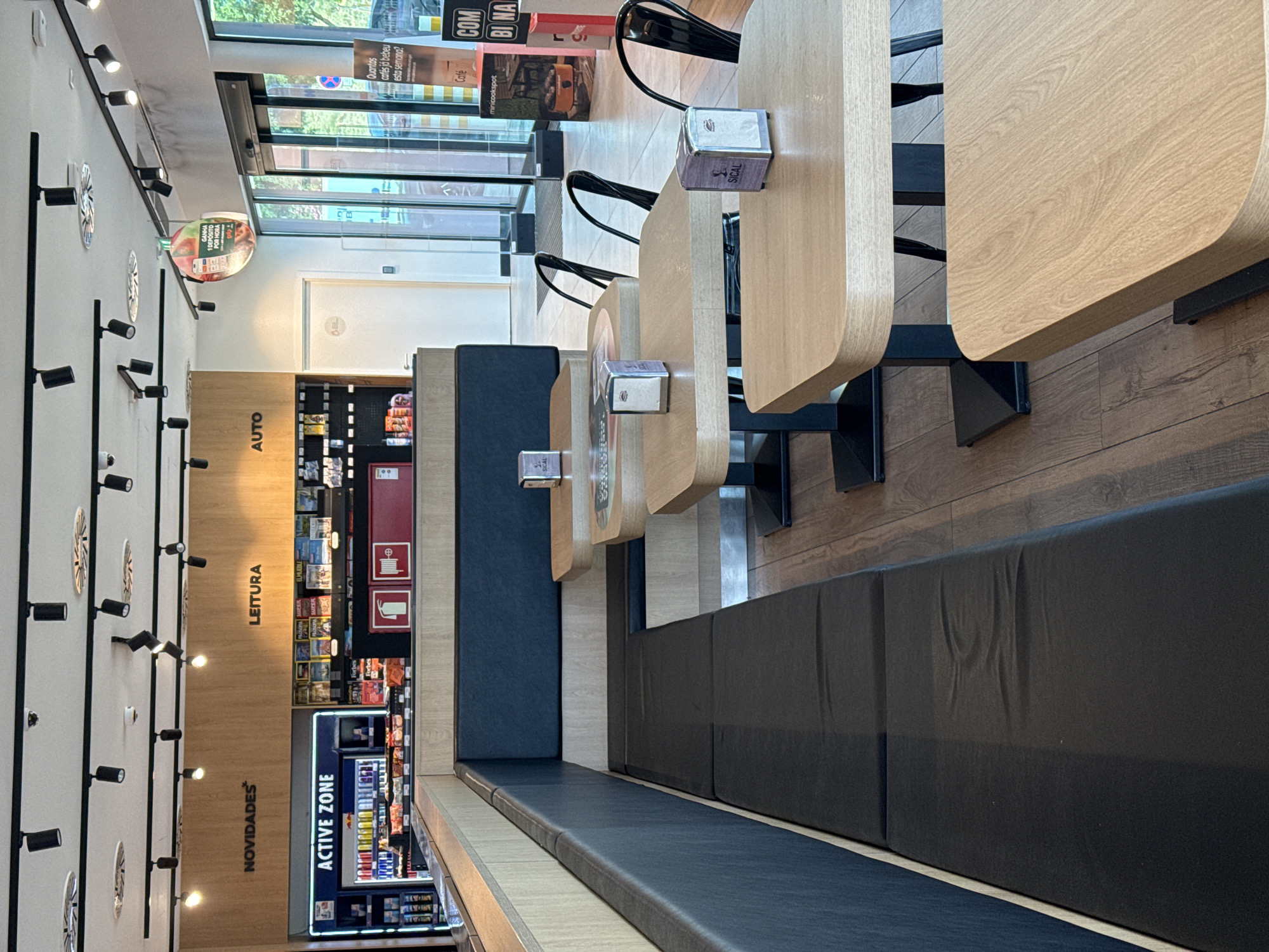 The image shows the interior of a service station shop in Oeiras, Portugal. The shop features a long, dark blue upholstered bench seat along the left side of the image, facing a row of light wood tables. Each table is set with a silver napkin dispenser. In the background, a counter area displays various products, including drinks in a refrigerator labeled "ACTIVE ZONE," magazines, and other convenience items. Above the counter, signs indicate "NOVIDADES," "LEITURA," and "AUTO." To the right, glass doors lead outside to a bright, sunlit area. A circular promotional sign hangs from the ceiling. The floor is made of dark wood planks. The overall impression is a clean and modern rest stop facility. There are no people visible in the image.