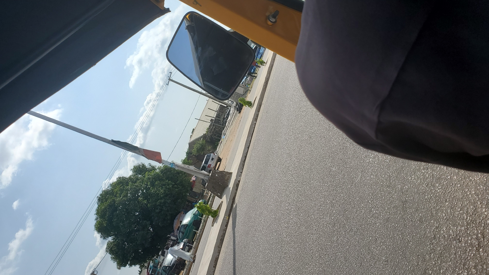 The image captures a street scene in Jalingo, Nigeria, viewed from the perspective of someone inside a yellow vehicle, likely a tricycle or tuk-tuk, indicated by the presence of a yellow-painted metal structure and a side mirror. The foreground shows a portion of the vehicle's dark-colored interior, possibly a seat or clothing, and the asphalt road.
In the mid-ground, a paved sidewalk runs parallel to the road. Several vehicles are parked and driving on the street, including a white SUV, a green car, and other unidentified cars. A man in white clothing stands near the sidewalk. A tall flagpole with a flag, partially unfurled and showing colors red, white, and green, stands prominently on the sidewalk. Trees and low-lying green bushes are visible along the sidewalk and in the background. Buildings with corrugated metal roofs line the far side of the street, suggesting a commercial or residential area. Telephone poles and overhead wires are present in the background, typical of an urban environment.
The sky is bright blue with scattered white clouds, indicating daylight, likely late morning or afternoon, given the light and shadows. The overall impression is a snapshot of everyday life on a street in Jalingo.