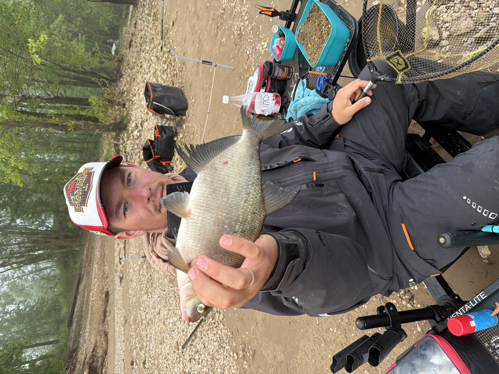 This image captures a man holding a freshly caught fish, likely a bream, on a pebbly river or lake bank in Chaikovsky, Russia. The man is seated, wearing a dark fishing jacket and dark pants, along with a white and red cap featuring "MAXIM" and "THE BEST SPINT RODS" text. A ring is visible on his left ring finger. He looks directly at the camera with a neutral expression, presenting his catch.
The background shows a line of trees obscured by mist or fog, indicating an overcast day. Around the man, various fishing accessories are set up: a blue container filled with groundbait and corn, other smaller containers for bait additives, and a clear plastic bottle with "КРАСНАЯ ЦЕНА" (Krasnaya Tsena - Red Price) and "СОК..." (Sok - Juice...) in Cyrillic. A blue towel, a fishing net with a fish logo, and rod holders are also visible, attached to what appears to be a fishing chair or station labeled "VENTA-LITE". Two black bags with orange accents are on the ground behind him. The scene portrays a calm moment during a fishing expedition.