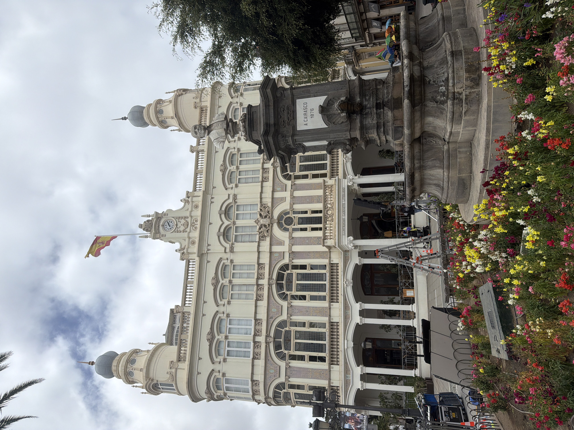 The image captures a wide-angle view of the Gabinete Literario building in Las Palmas de Gran Canaria, Spain, on an overcast day. The ornate, cream-colored building features classical architectural elements, including arched windows, decorative molding, and two distinctive domed towers. A Spanish flag flies prominently from the rooftop. In the foreground, a large, dark stone monument with a bust of a man stands beside a flower bed filled with a vibrant array of red, yellow, white, and pink flowers. To the right of the monument, a large green tree provides shade. The area in front of the building appears to be undergoing some form of setup or activity, with several ladders, scaffolding, and what look like film or event equipment present. A banner with Spanish text is visible on a lamppost to the left. Several people are casually dressed and appear to be either working or observing the scene. The overall impression is one of a public space in front of a significant cultural building, with evidence of recent or ongoing event preparation.