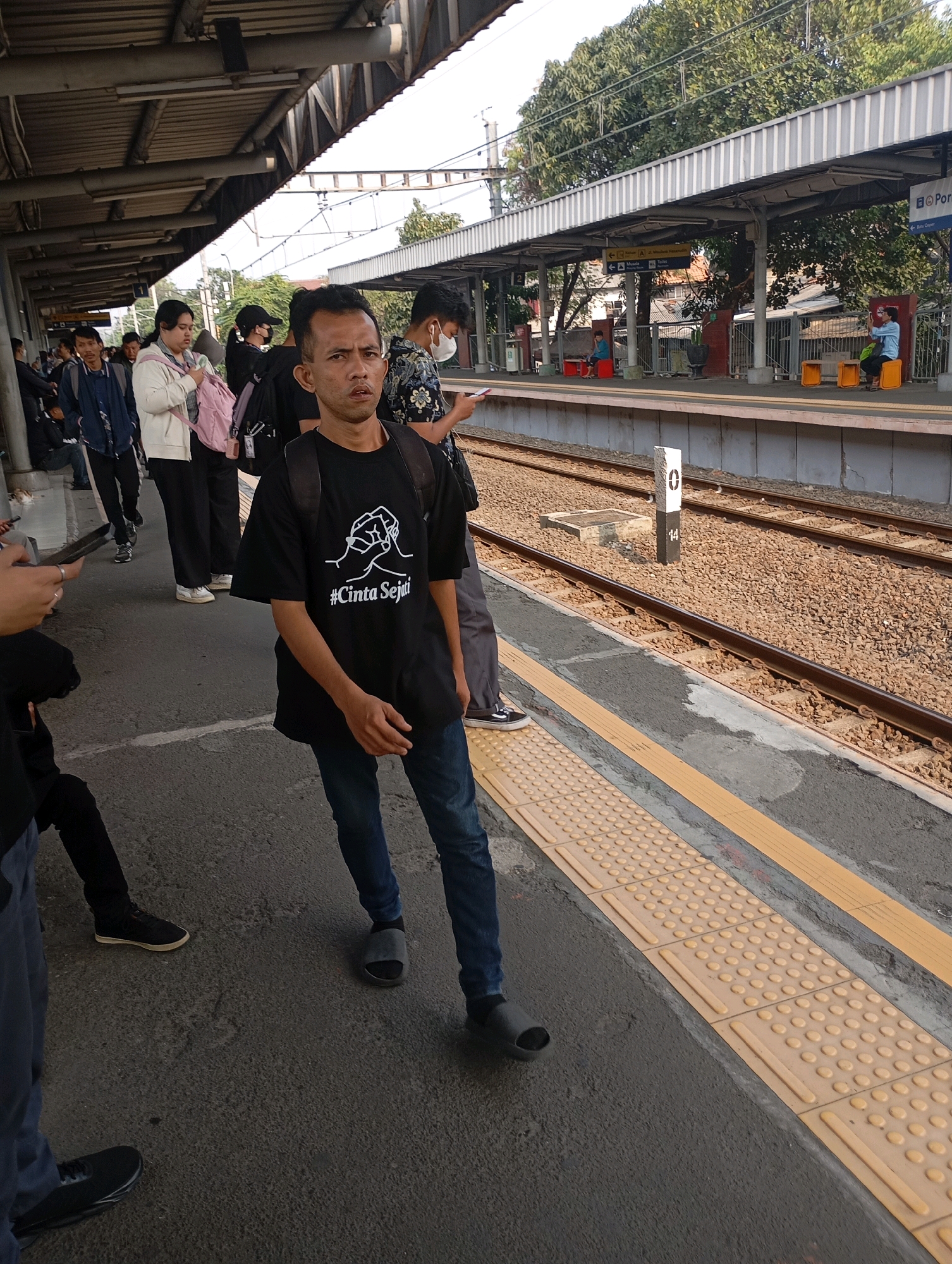 The image depicts a busy train station platform in Tangerang, Indonesia. In the foreground, a man wearing a black t-shirt with a white graphic and the text "#Cinta Sejati" (True Love in Indonesian) and blue jeans stands looking towards the camera. He has a backpack on. Several other individuals are visible on the platform, some appearing to be waiting for a train. The platform features tactile paving for the visually impaired, indicating accessibility measures. Train tracks and overhead electrical wires are present, typical of a railway environment. The overall scene suggests an active public space during daylight hours.