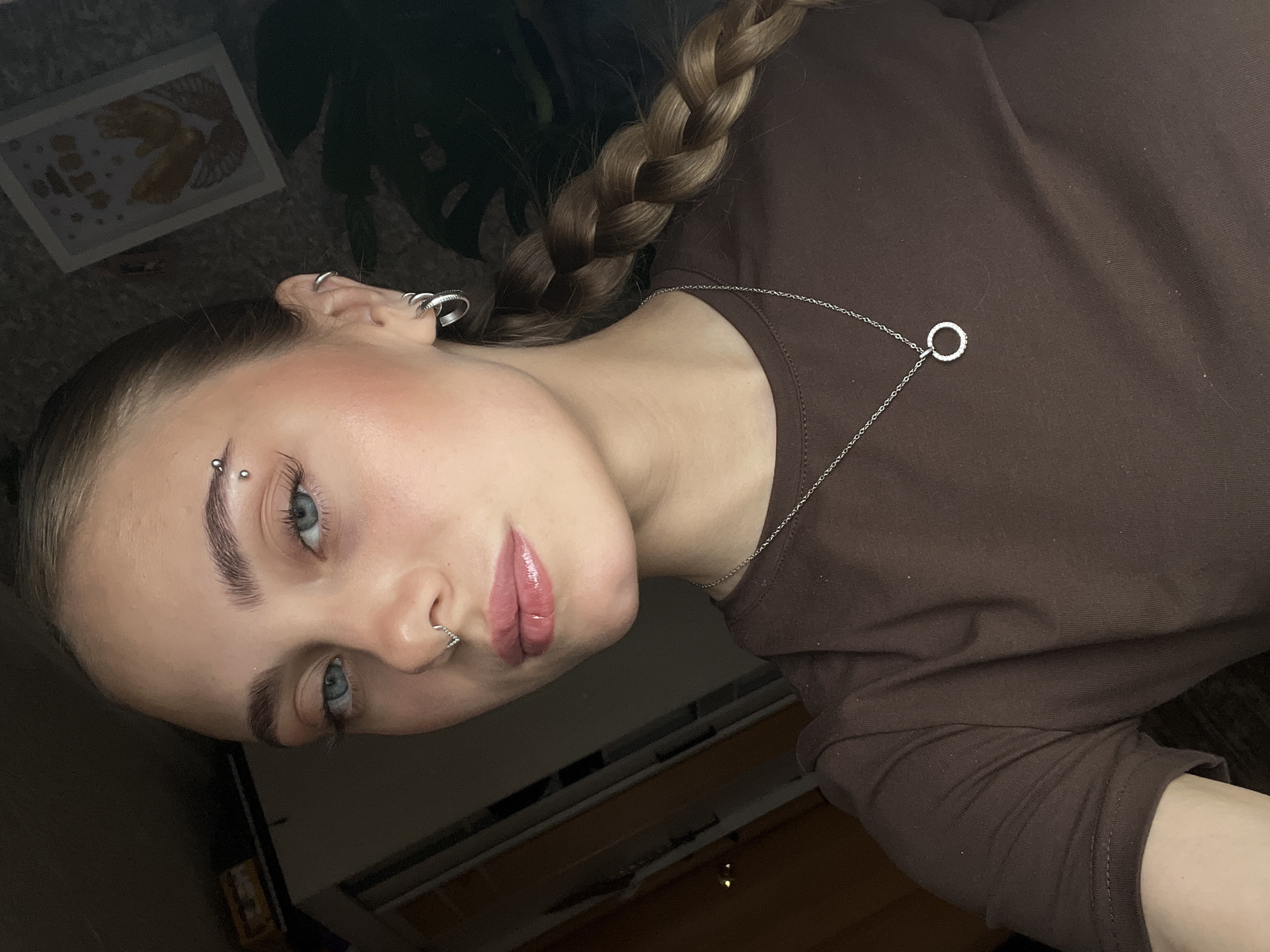 An angled portrait captures a young woman with fair skin, light blue eyes, and light brown hair braided over her left shoulder. She looks directly at the camera with a calm, composed expression. Her face is accented with makeup, including a soft pink blush on her cheekbones and glossy pink lips.
She has multiple piercings: a curved silver barbell on her left eyebrow, a heart-shaped silver hoop in her septum, and several silver hoops and studs adorning her left ear. She is wearing a dark brown, long-sleeved top. A thin silver chain necklace with a circular, possibly stone-encrusted, pendant hangs around her neck.
The background is dark and largely out of focus, indicating an indoor setting under low light conditions. In the upper left, a framed piece of artwork is partially visible, depicting a light-colored, abstract or winged figure. Dark foliage from an unseen plant is discernible in the upper right. The image appears to be a posed portrait or selfie. There is no visible text within the image. The provided location context is Фоки, Russia.