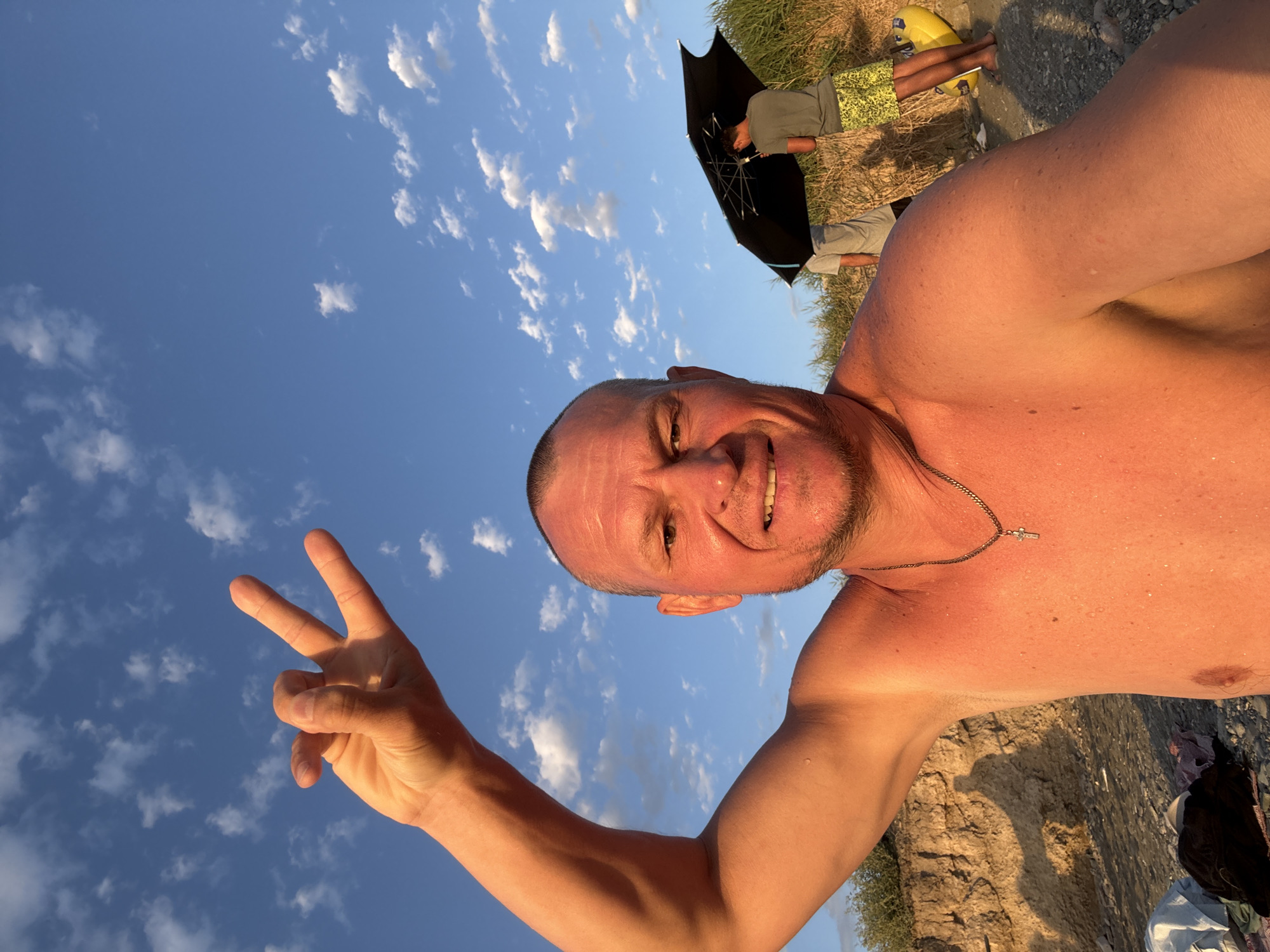 In Zharbulak, Kazakhstan, a man in his 40s, with a shaved head and a silver chain around his neck, smiles at the camera, holding up his right hand with his thumb and index finger extended, forming a 'V' shape against a bright blue sky dotted with white clouds. The setting sun casts a warm, orange glow on his bare chest and arm, highlighting sweat droplets. In the background, two other individuals are visible; one holding a black umbrella, and another wearing green patterned shorts. The ground is rocky with some vegetation and a yellow inflatable object. The overall scene depicts a casual outdoor gathering by a body of water, likely at sunset.