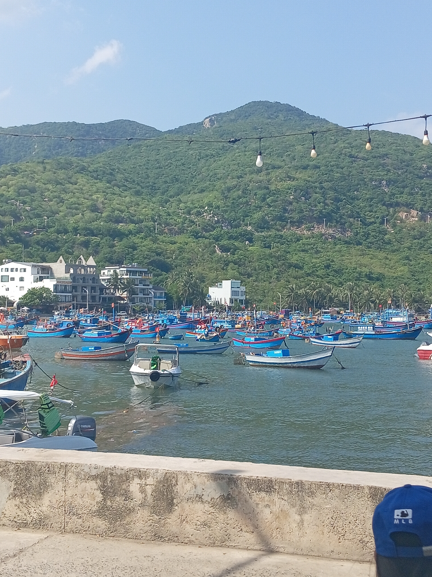 The image shows a harbor filled with numerous blue and white fishing boats, docked near a shore with buildings and a lush green mountain in the background. The sky is clear and blue with a few scattered clouds. In the foreground, a concrete wall runs across the bottom of the frame, with a portion of a blue baseball cap visible on the right side, bearing an MLB logo. The scene is consistent with the coastal area of Vĩnh Hải, Vietnam, known for its fishing industry and scenic mountainous backdrop.