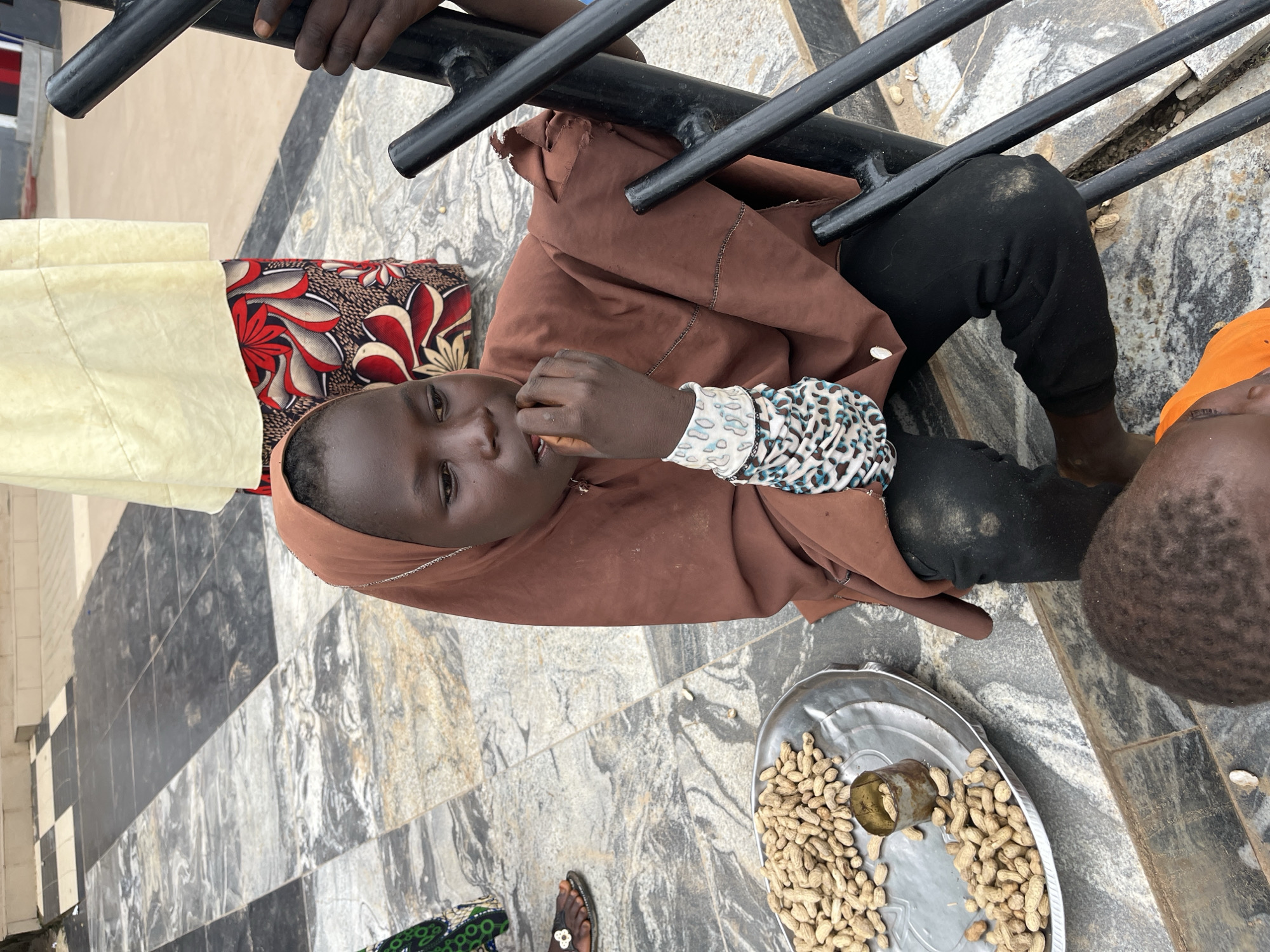 In Galadimawa Village, Nigeria, a young person wearing a brown hijab and dark clothing sits on stone steps, eating peanuts from a metal tray. The tray contains a pile of peanuts and a small, rusty tin. The person's right hand is in their mouth, and their left arm is adorned with patterned bracelets. To the left of the tray, a person's foot in a sandal is visible. In the background, another person stands wearing a pale yellow garment over a patterned skirt. A black metal railing is positioned to the right of the seated individual. The ground is paved with marbled tiles.