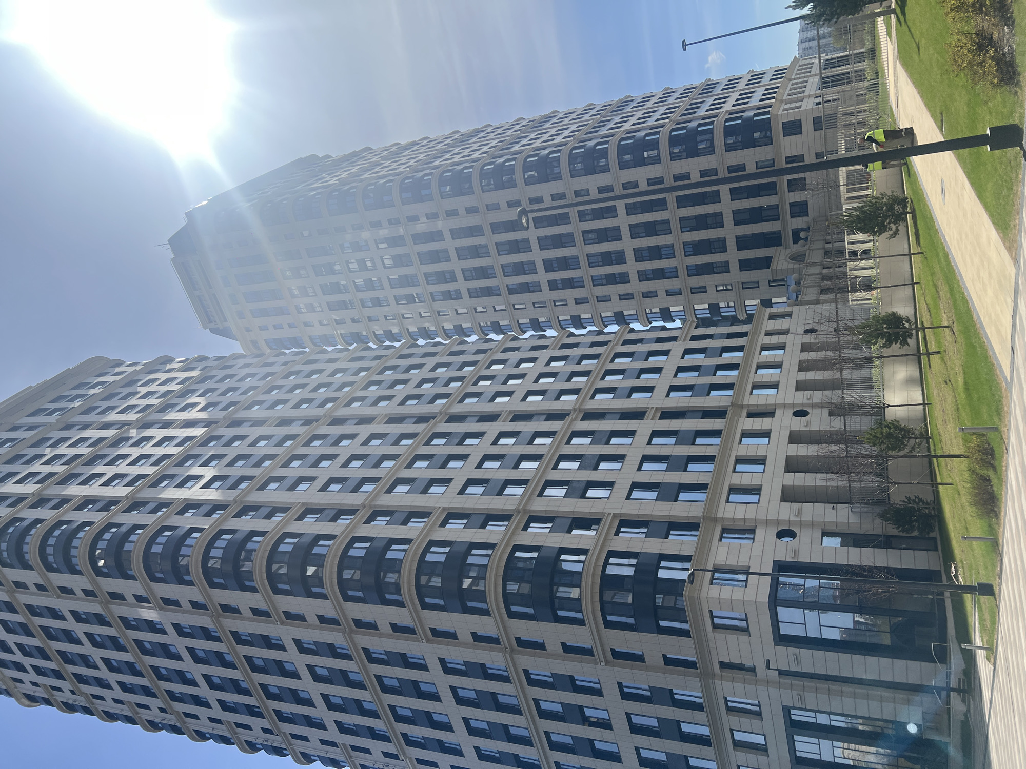 This image, captured on a sunny, clear day, depicts two modern, tall high-rise buildings with a distinctive architectural style. The buildings feature light-colored facades contrasted with dark window frames, incorporating a repetitive pattern of rectangular windows and sections with curved, dark-framed windows or balconies. Bright sunlight, emanating from the upper left, casts glare across the top of the structures.
The buildings are situated in a well-maintained outdoor setting, featuring paved pathways, neatly manicured grassy areas, and several young trees. Along one of the pathways, a person wearing a yellow or green high-visibility vest and dark pants is visible, appearing to be a maintenance or cleaning worker, possibly pushing a cart. Modern streetlights are also present along the path.
No discernible text is visible on the buildings or any other objects. The scene suggests a contemporary residential or commercial complex, possibly located in an urban or suburban area like Khimki, Russia. The image is presented with a significant tilt, making the buildings appear to lean.