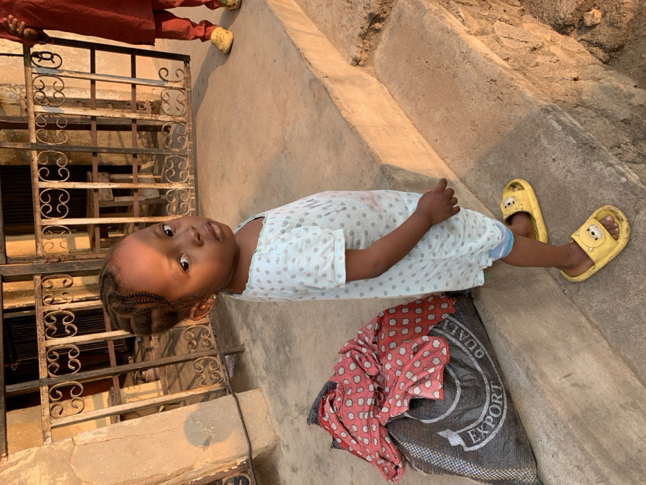 A young child wearing a light blue patterned dress lies on a concrete surface next to a patterned cloth and a black bag. The child has braided hair and wears yellow cartoon-character sandals. A metal gate sits to the side. Part of another person's legs in reddish-brown clothing and yellow sandals is also visible in the top left corner.
