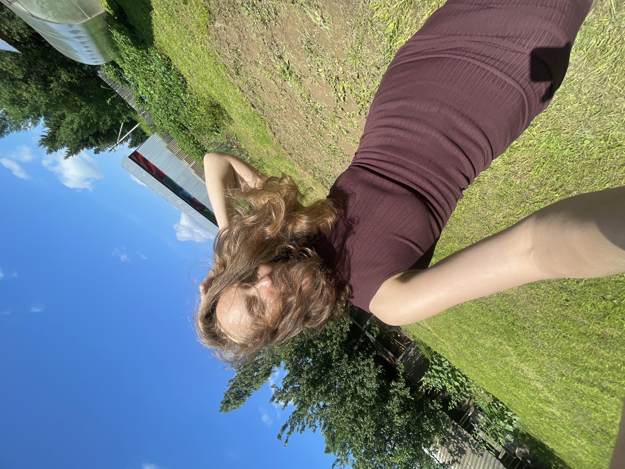 A young woman with light brown hair is captured in a selfie outdoors, likely in the village of Sharkan, Russia, given the context. She is wearing a dark red, ribbed dress and her hair is being blown by the wind. The background features a clear blue sky with scattered clouds, lush green grass, a tilled garden patch, trees, a wooden fence, and a long, silver-colored structure that appears to be a greenhouse or similar agricultural building. A portion of a house with a corrugated roof is also visible on the left side of the image. The woman is standing in an open area, possibly a backyard or a field.