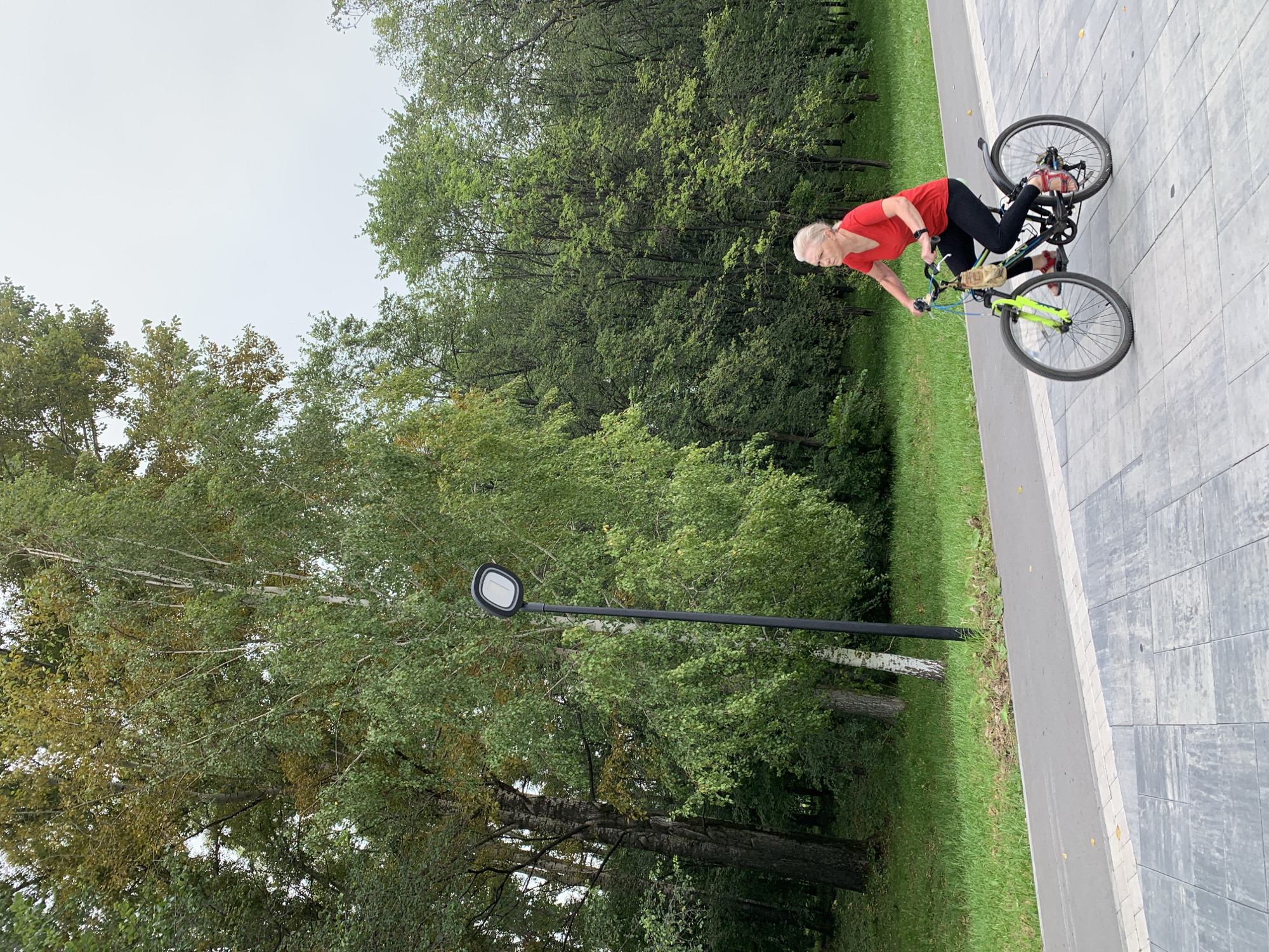 The image depicts an elderly woman with blonde hair, wearing a red t-shirt and black leggings, cycling on a paved path. She is on a bicycle with a neon green front fork and black frame. The background consists of lush green trees and grass, suggesting an outdoor park setting. A lamppost is visible to the left of the woman. The sky is overcast and appears pale. The paving stones in the foreground are grey and textured. The scene is likely from Prokopyevsk, Russia, given the context.