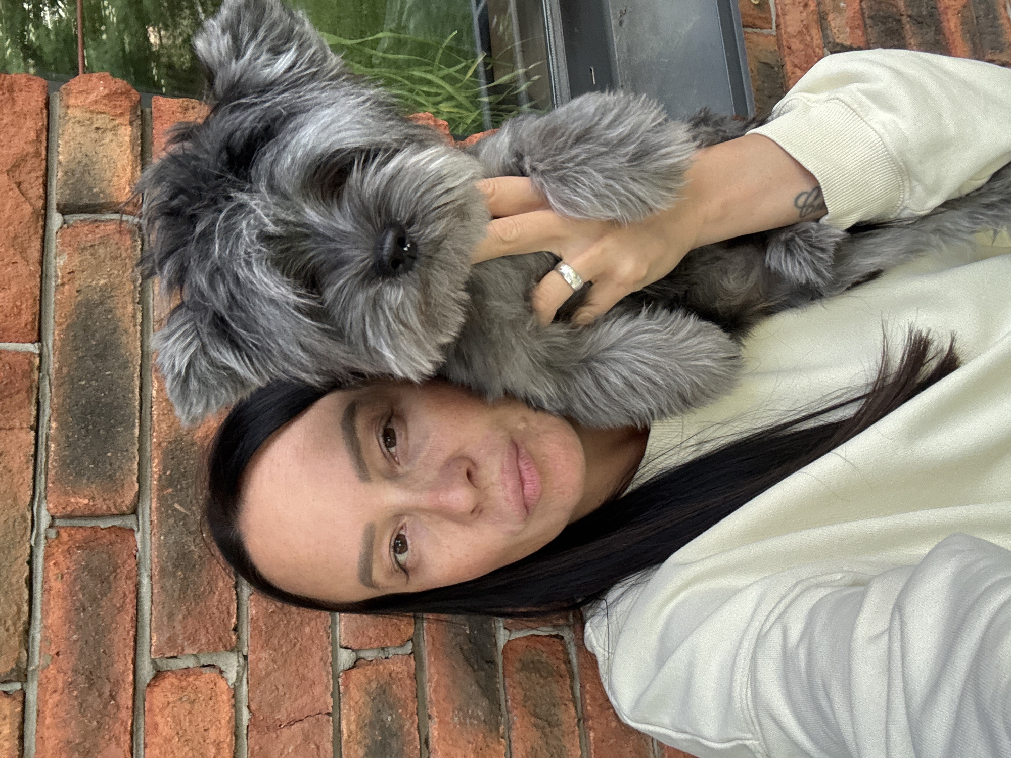 A woman with dark hair is lying on a brick surface, with a gray, furry dog nestled on her chest and held by her hands. The woman is wearing a white sweatshirt. A silver ring is visible on her right hand. In the background, there's a glimpse of greenery and a window or reflective surface. The location cannot be determined from the image provided.