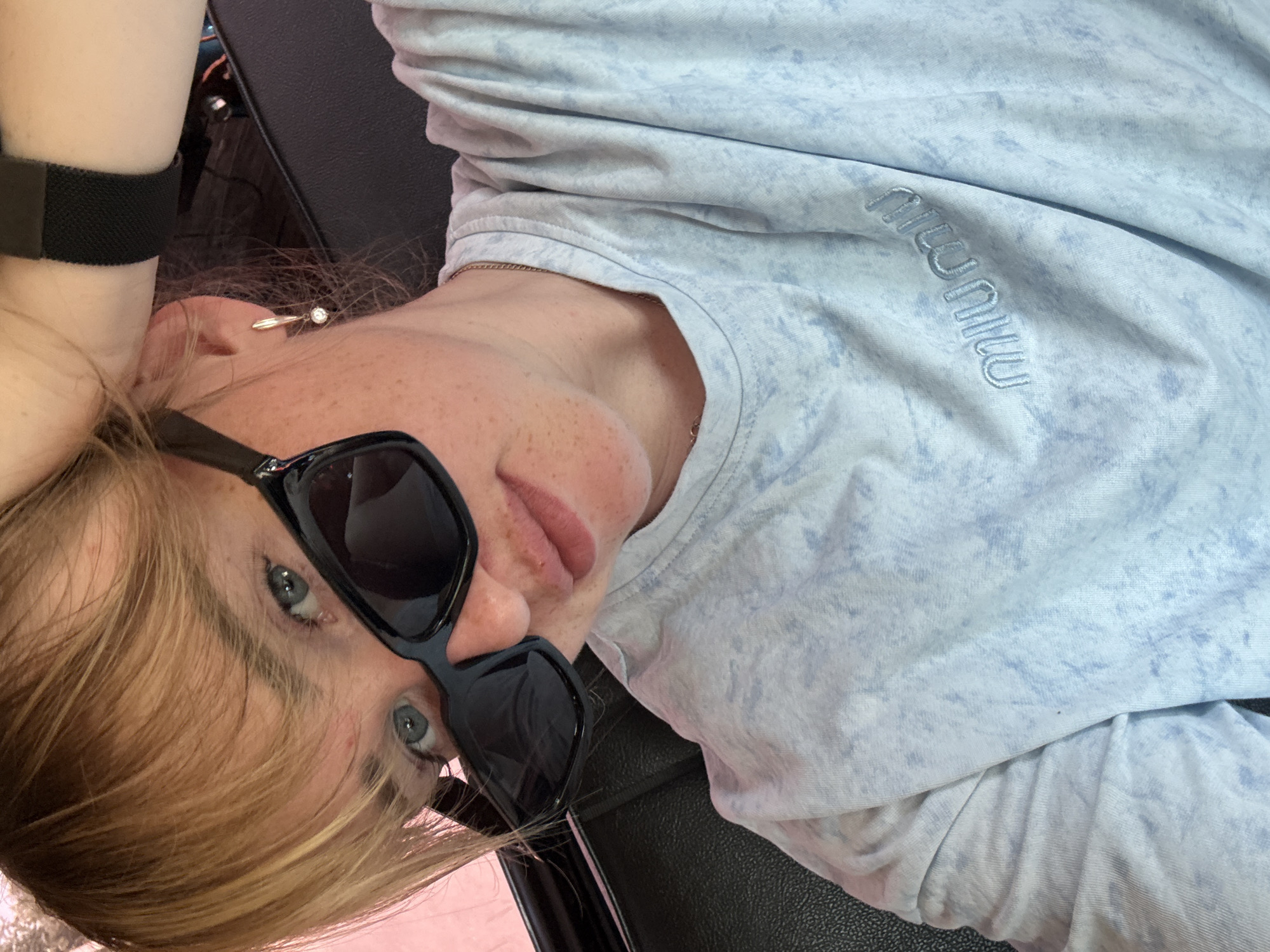 A young woman with blonde hair, wearing sunglasses and a light blue patterned t-shirt, is looking at the camera. She has freckles on her face and is wearing a gold earring. Her left hand is raised and resting on the back of her head. The background appears to be the interior of a vehicle, possibly a car, with dark leather seats visible. The image does not provide any specific information about the location being Chaikovsky, Russia.