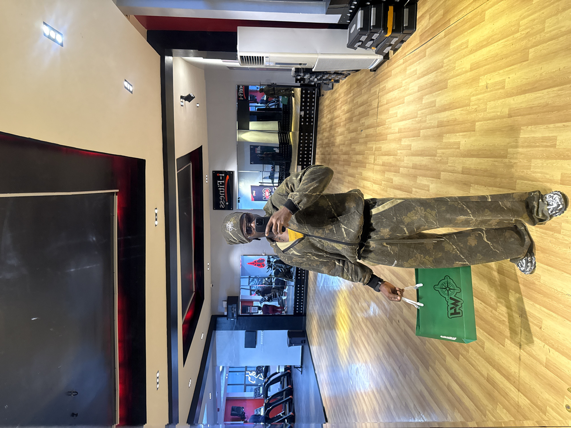 A man stands in a gym, taking a selfie in a mirror. He is wearing a camouflage tracksuit and a beanie, and holding a green shopping bag. The gym is well-lit, with wooden floors, treadmills, and other exercise equipment visible. A sign in the mirror reads "I-FITNESS". The man appears to be in his late 20s or early 30s. The gym is located in Lekki, Nigeria.