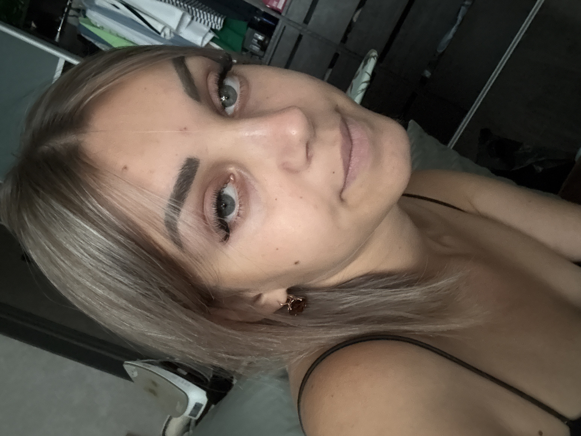 A young woman with blonde hair is posing for a selfie in what appears to be a bedroom. She is wearing a black camisole and a brown earring. Behind her, a closet full of clothes is visible, along with a dresser. The woman is looking directly at the camera with a slight smile. The lighting in the room is dim. The scene does not provide any specific indicators that would allow for the determination of its location within the city of Yuzhno-Sakhalinsk, Russia.