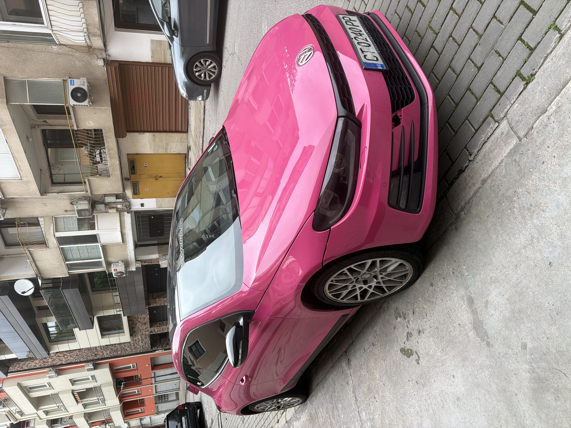 An overhead, slightly tilted shot captures a bright pink Volkswagen parked on a paved street in what appears to be an urban setting. The car, possibly a Scirocco model given its design and the VW badge, is the central subject, its vibrant color contrasting with the muted tones of the street and buildings. It features black accents on the grille and headlights, and multi-spoke silver alloy wheels.
Behind the pink car, a gray hatchback is partially visible. The street surface is a mix of gray interlocking brick pavers with some moss growth and rough concrete.
The background consists of multi-story residential or mixed-use buildings, characteristic of a European city, specifically Plovdiv, Bulgaria, as indicated by the license plate. The architecture varies, with some buildings having rough stucco facades, balconies with metal railings, and numerous air conditioning units affixed to their exteriors. One building features dark brown roll-down shutters over a ground-floor entrance, and a yellow double door below it. Another building has a modern facade with dark panels and multiple windows. A satellite dish is mounted on one of the buildings.
The scene takes place during the daytime, under what appears to be an overcast sky, as indicated by the soft, diffused lighting and lack of harsh shadows. There are no people visible, and the cars appear to be stationary.
Visible text includes the Bulgarian license plate "CT 0240 PP" on the pink car, with "BG" (Bulgaria) in a blue strip on the left. "CT" is the vehicle code for the Plovdiv district, confirming the location. A sticker on the car's rear side window also reads "VWclub.bg".