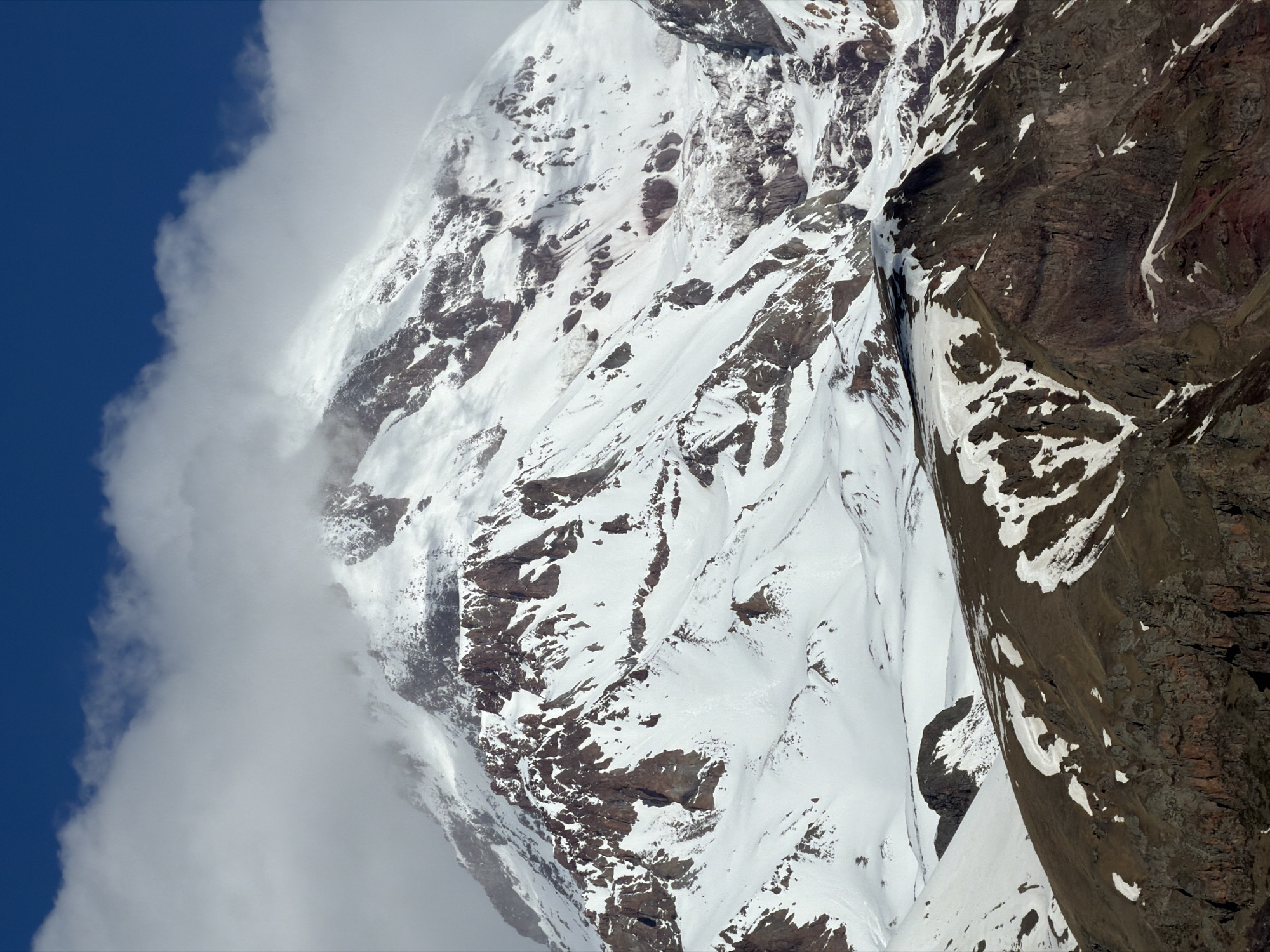 The image depicts Mount Kazbek in Stepantsminda, Georgia. The mountain face is partially covered in snow and interspersed with rocky terrain. A thick cloud bank obscures the left side of the mountain, contrasting with the blue sky in the upper-left corner.