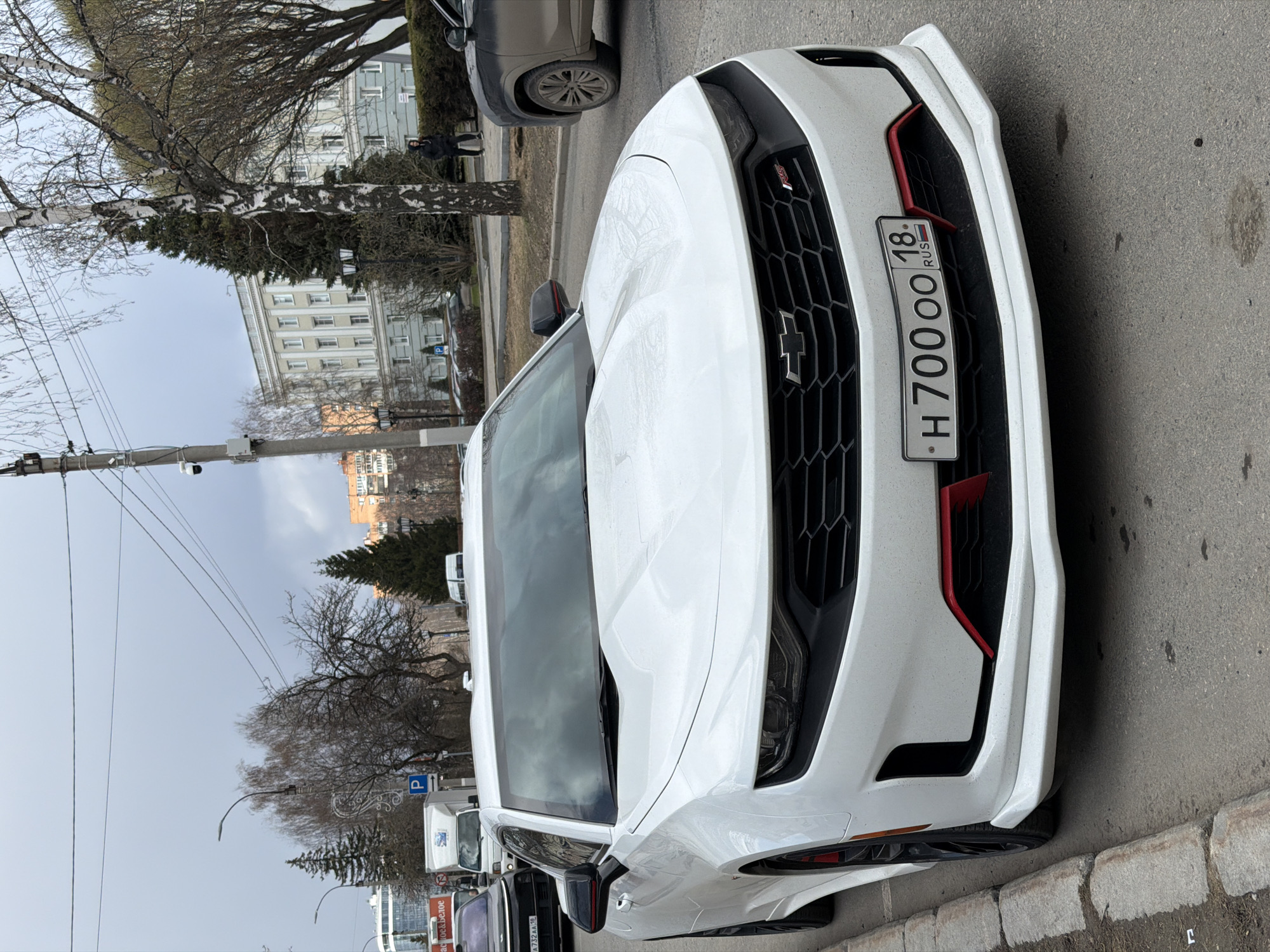 A white Chevrolet Camaro SS is prominently featured, parked diagonally on the side of a city street. The car's front is angled towards the viewer, displaying a black hexagonal grille with a Chevrolet emblem and red decorative accents on the bumper. Its Russian license plate, "H 700 OO 18 RUS," is clearly legible, with "18" indicating the Udmurt Republic, where Izhevsk is located. The car appears slightly dirty, suggesting recent travel or a dusty environment.
The setting is an urban street scene in Izhevsk, Russia, under an overcast, grey sky. The weather appears cool, with bare deciduous trees, including a large birch tree with white bark and hints of snow or ice, alongside evergreen foliage. In the background, a large, multi-story building with many windows dominates, and another building with an orange-brown facade is visible further away. A dark grey SUV is parked behind the Camaro, and other vehicles, including a white truck, line the street. A "P" parking sign is visible.
One person, dressed in a dark jacket, stands in the background near the birch tree. Utility poles and overhead wires crisscross the upper left portion of the image. Visible text on signs includes "Пятерочка & Белое" (Pyaterochka & Beloe), referring to Russian retail chains. The scene captures a quiet, ordinary moment on a city street, likely in late winter or early spring, with no specific activity beyond parked vehicles.