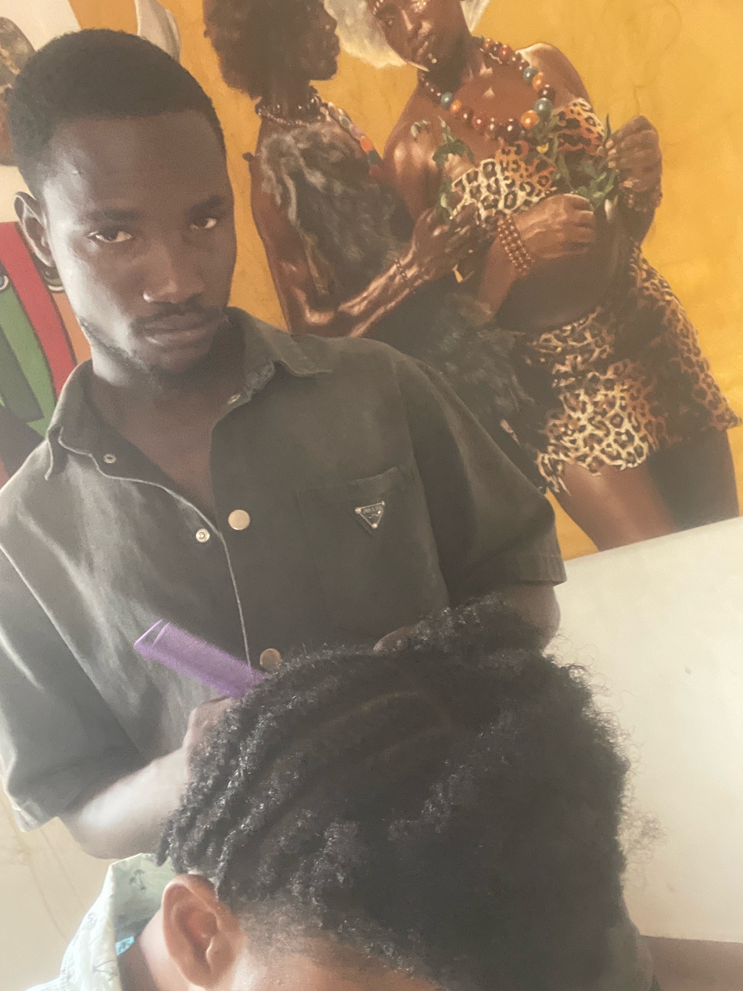 In Ado, Nigeria, a man with dark skin and short hair is seen from the chest up, facing the camera. He is wearing a dark green collared shirt with buttons. In front of him, another person's head is visible from the side, also with dark skin and textured hair. The second person's hair is being styled with a purple comb by the man in the foreground. Behind them, a mural on a yellow wall depicts two figures with traditional African attire, including leopard print clothing and beaded necklaces. One figure holds a plant. The overall scene appears to be an indoor setting, possibly a salon or home.