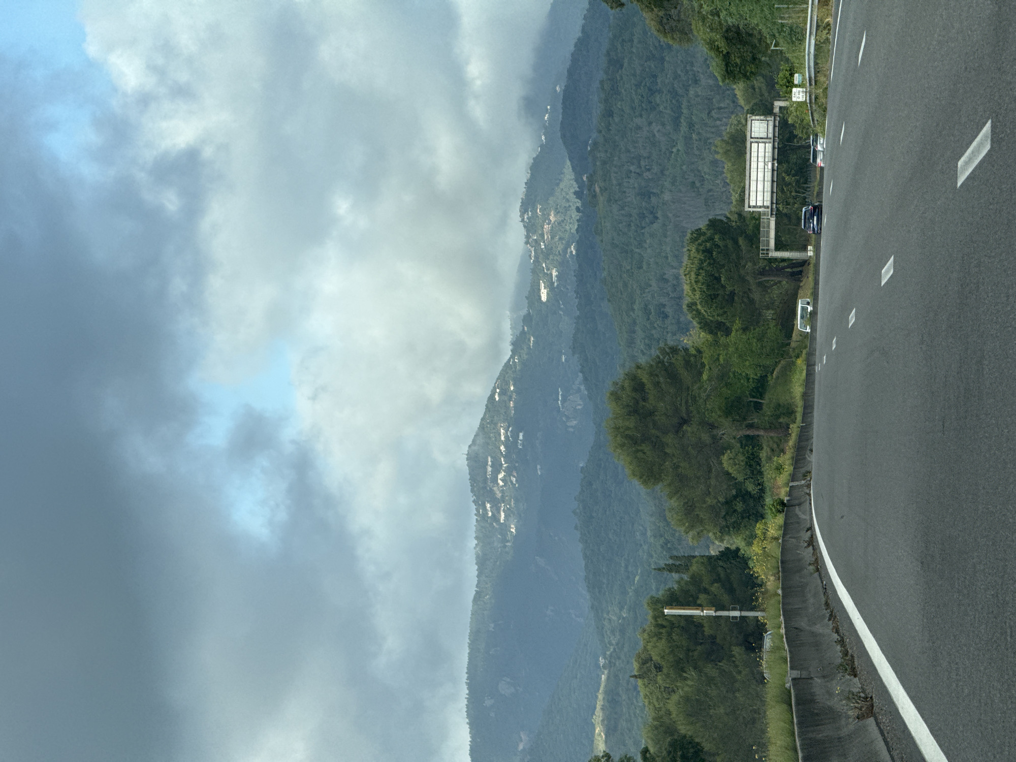 The image shows a highway winding through the mountainous terrain of Mandelieu-la-Napoule, France. The road occupies the right side of the frame and is bordered by a white line. Mountains covered in dense green vegetation rise on the left side, their peaks partially obscured by heavy, overcast clouds. Houses or buildings can be seen scattered on the slopes of the mountain. A highway sign is visible along the side of the road. A few vehicles are present on the highway.