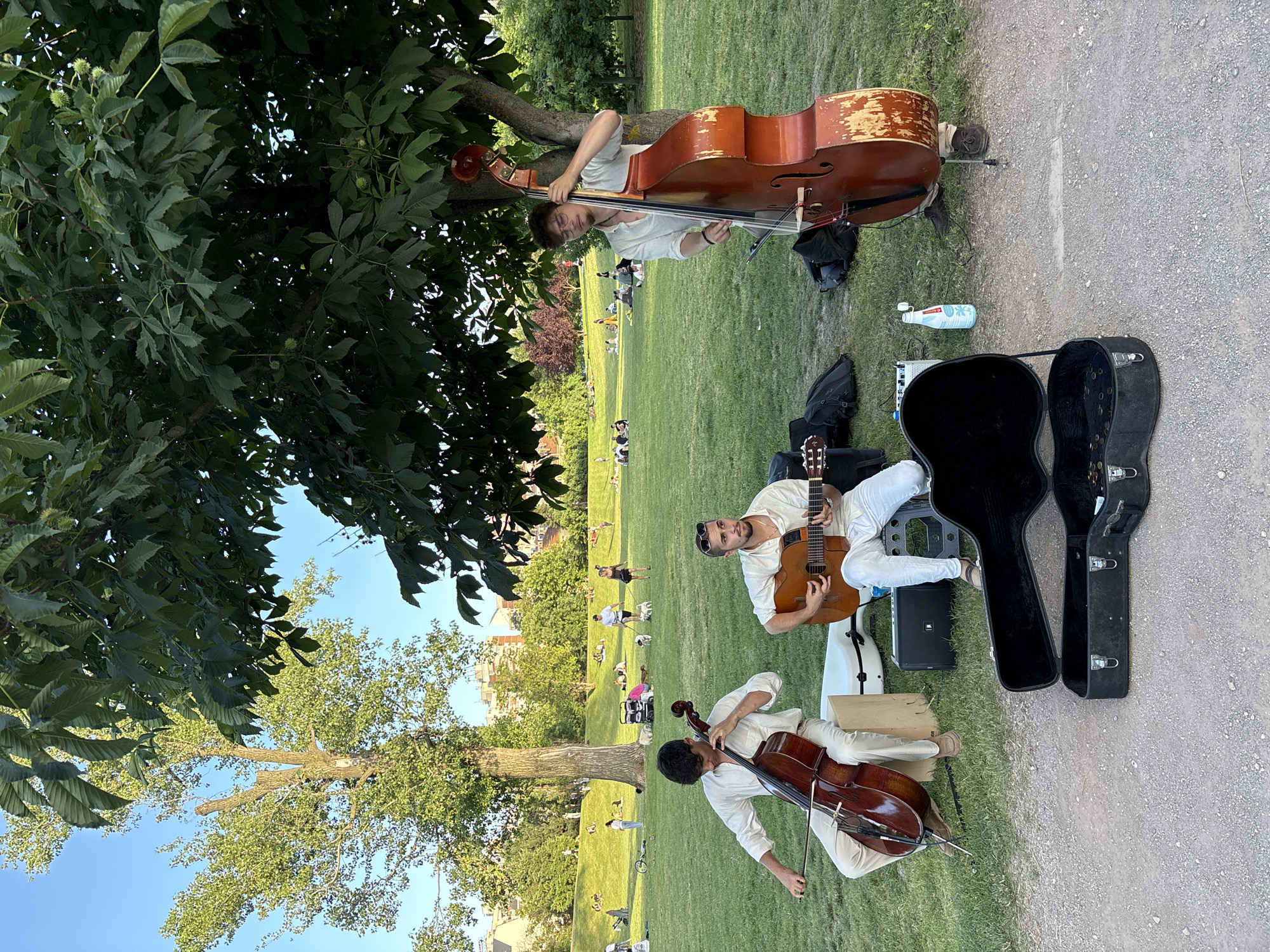 Three men are playing music in a park on a sunny day. The man on the left is playing a cello, the man in the middle is playing a guitar, and the man on the right is playing a double bass. They are all wearing white clothing. In the background, there are people relaxing and walking in the park. The park appears to be a large, grassy area with trees and buildings in the distance. The image is taken from a slightly low angle, looking up at the musicians. The ground in the foreground is covered with gravel, and there are instrument cases and a small speaker. The location is a park in Sofia, Bulgaria.