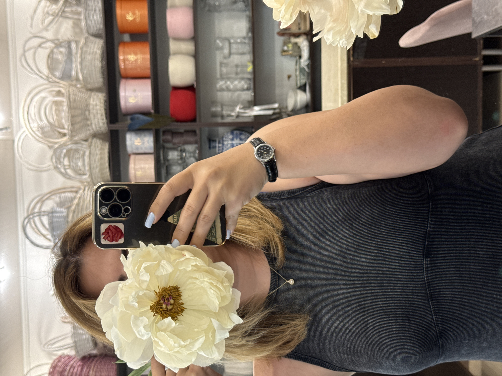A person holds a smartphone up to a mirror, taking a selfie. The person is wearing a dark grey tank top and a watch with a black band on their left wrist. A white flower, possibly a peony, is held in front of the person's face, partially obscuring it. The background is a reflection of shelves filled with various items, including small woven baskets, cylindrical containers in shades of orange, pink, and red, and some glassware. The overall scene appears to be indoors, possibly in a shop or storage area. The lighting is consistent with indoor lighting. There is no visible text in the image. The activity is a photo-taking moment.