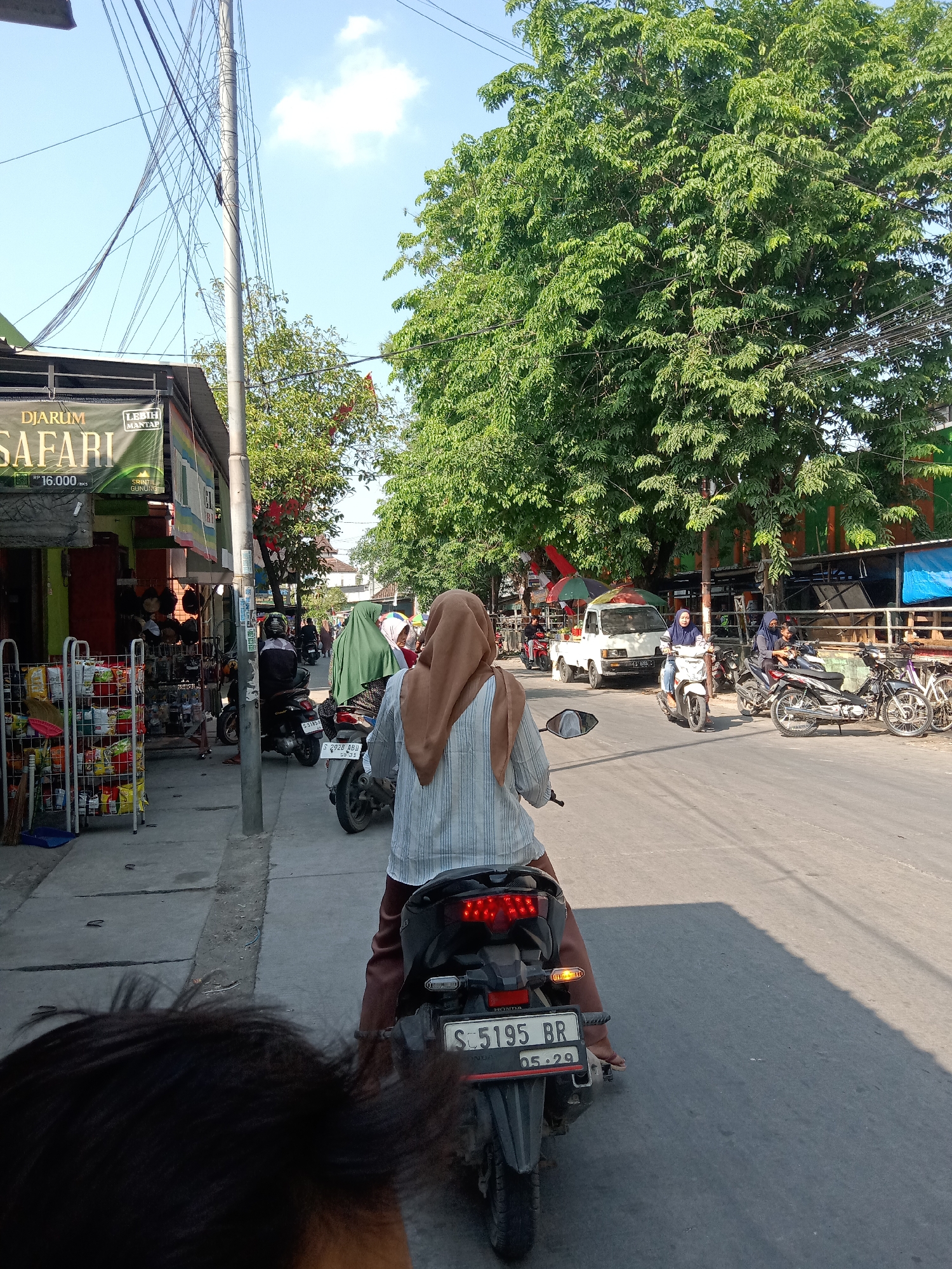 The image shows a street scene in Sugihwaras, Indonesia. The perspective is from behind a person on a motorcycle, looking forward. The street is paved and has a sidewalk on the left. Several motorcycles are visible, some parked and some being ridden. On the sidewalk, there are shops with merchandise displayed outside. People are seen riding motorcycles and walking. Trees line the right side of the street, providing shade. The sky is blue with some clouds. The overall atmosphere suggests a busy, everyday street in a tropical urban environment. The license plate of the motorcycle in the foreground reads "S 5195 BR".