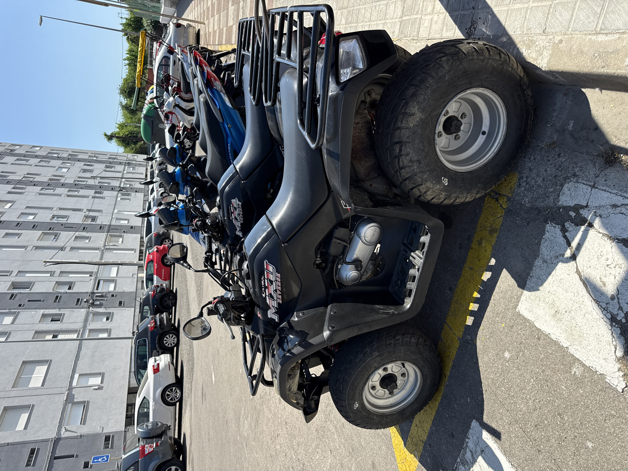 A black all-terrain vehicle (ATV) is parked on asphalt pavement in front of a tall, grey apartment building. The ATV is in the foreground, angled slightly to the left, with its front left wheel visible. The vehicle has a black seat and black racks on the front and rear. Yellow and white painted lines are visible on the asphalt. Behind the ATV, several motorcycles are parked in a row. In the background, the apartment building has many windows. The sky is clear and blue. The scene appears to be in a city, likely Blanes, Spain, given the architectural style and the presence of parked vehicles typical of Mediterranean urban areas.