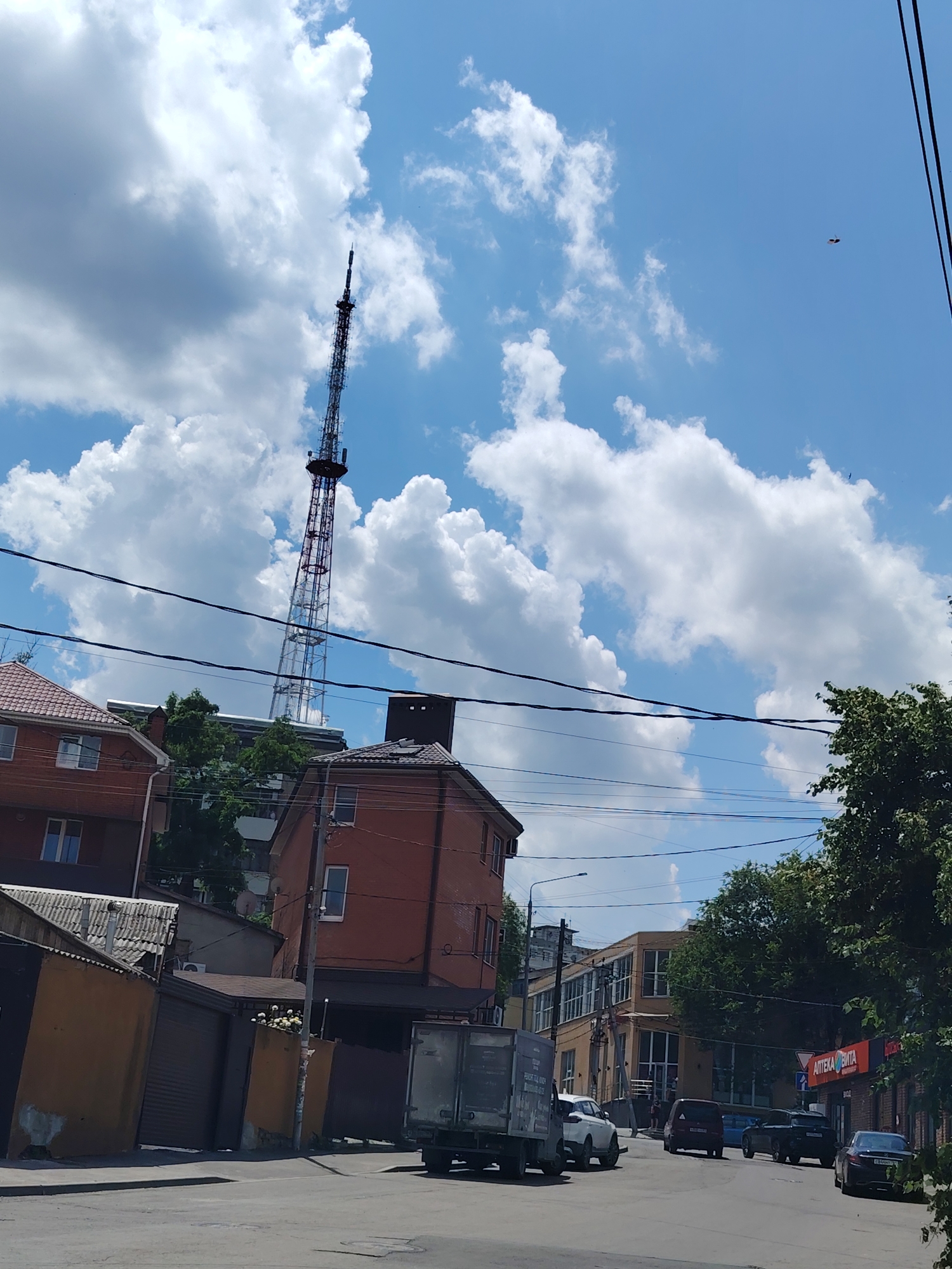 The image is a street-level view of an urban environment. Dominating the background is a tall, lattice-style transmission tower, likely a television or radio mast, set against a bright blue sky with scattered white clouds. In the midground, several buildings are visible. These include multi-story residential or commercial buildings made of brick, with pitched roofs and windows. Some appear to be older, while others look more modern. A street with several parked and moving vehicles is in the foreground and midground. The vehicles include cars, vans, and a truck. Some of the buildings have signs, one of which reads "АПТЕКА ВИТА" (Pharmacy VITA) in Cyrillic. Trees line parts of the street, and overhead power lines crisscross the scene. The overall impression is of a bustling city street on a clear day. The presence of the "АПТЕКА ВИТА" sign and the Cyrillic script strongly suggests the location is in Russia, and given the specific appearance of the transmission tower, it can be identified as the Rostov-on-Don television tower, located in Rostov-on-Don, Russia.