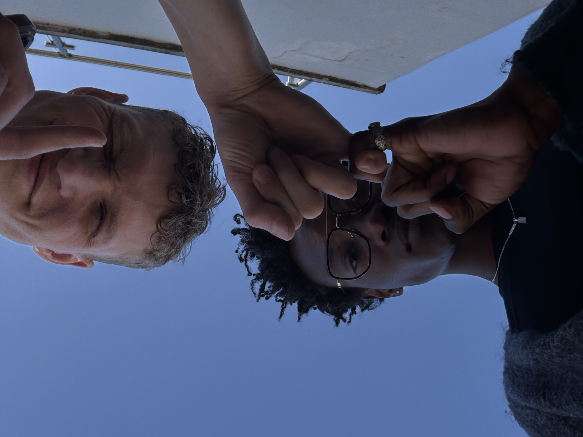 The image, taken from a low angle in London, United Kingdom, captures two individuals looking down towards the camera against a clear blue sky.
On the left, a person with a lighter skin tone and short, curly light brown hair is visible. They are making a "V" or peace sign with their left hand, held near their face. A ring is present on one of their fingers.
On the right, another person with a darker skin tone and dark, curly hair, possibly dreadlocks, is shown. This individual wears glasses with thin frames and a silver-colored necklace featuring a rectangular pendant. A prominent ring is also visible on a finger of their right hand.
The hands of both individuals are centrally placed and appear to be interacting or held closely together, with the left person's hand overlapping the right person's hand.
At the very top of the frame, a light-colored structure, possibly an awning or the underside of a ceiling, provides a partial architectural element. The predominant background is a bright, clear blue sky, indicating a sunny daytime setting. The overall impression is a casual and relaxed interaction. There is no visible text in the image.