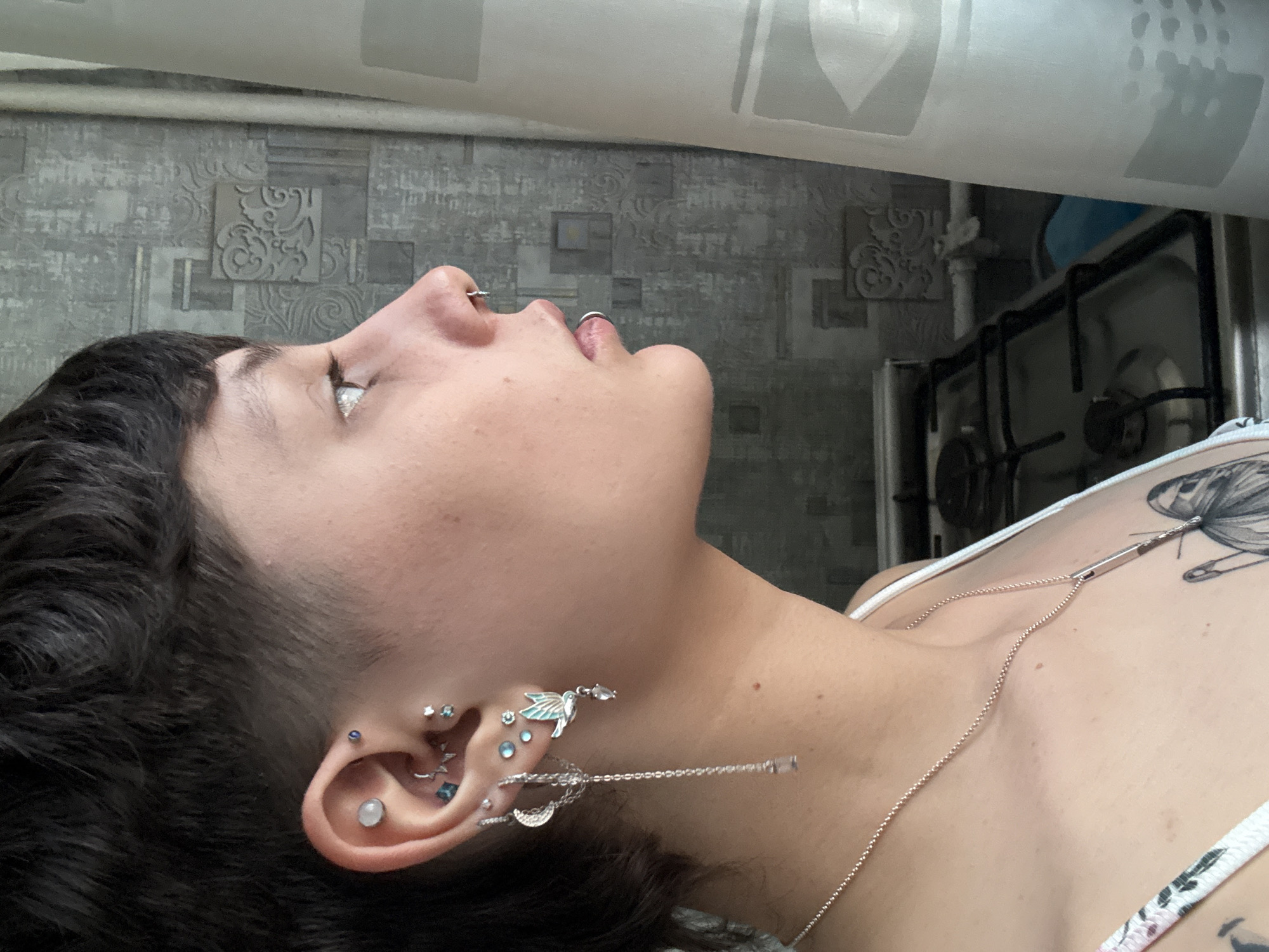 A close-up, angled shot features a woman from the chest up. Her head is tilted back, and her face is turned to the right side of the frame. She has dark hair, cropped short on one side, revealing multiple ear piercings adorned with various earrings including a dangling earring with a moon, leaves, and a tear drop stone. She has a ring in her nose and her lips are slightly parted. She is wearing a white tank top with a floral pattern on the straps, and a silver chain necklace that runs diagonally across her chest. The background includes a patterned grey wall and a glimpse of a metal stove or oven unit. There is insufficient information in the image to determine the location in Saint Petersburg, Russia.