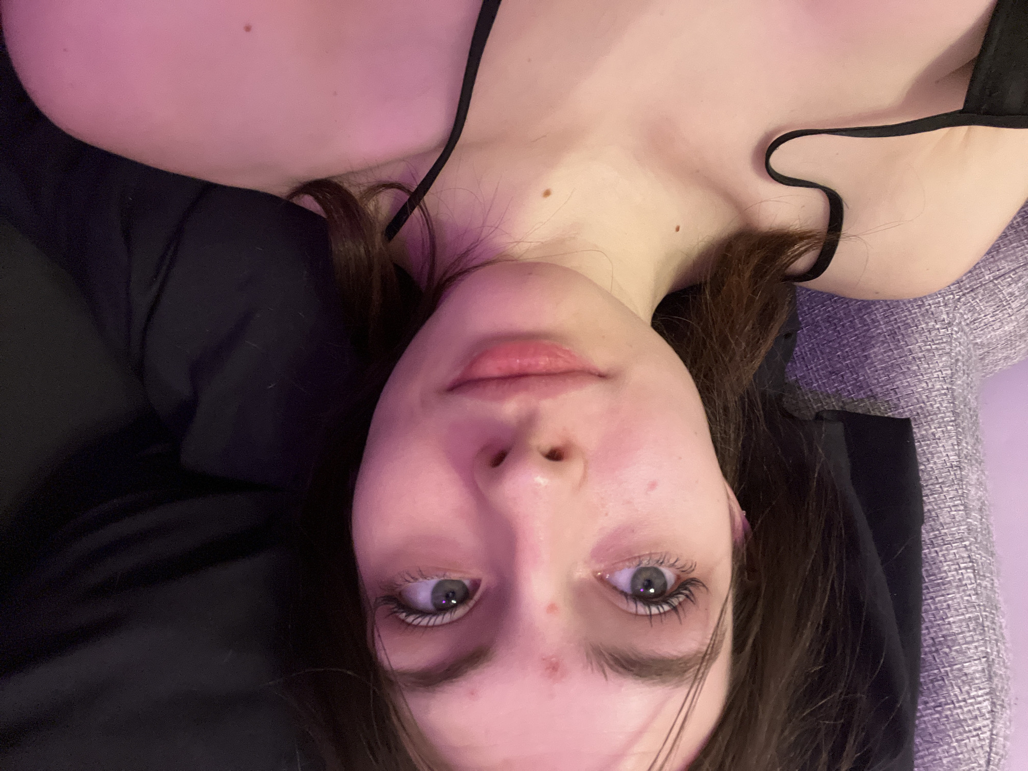 A close-up, upside-down view captures a young woman with fair skin and dark brown hair. She is lying down, with part of her upper chest and shoulders visible. She appears to be wearing a black top with thin straps. Her eyes are a pale blue-gray, and she has some minor blemishes on her face. The background includes a dark, solid material, likely clothing or a blanket, and a textured, gray-toned surface that could be upholstery. There is nothing in the image to indicate a specific location in Saint Petersburg, Russia, or any other city.
