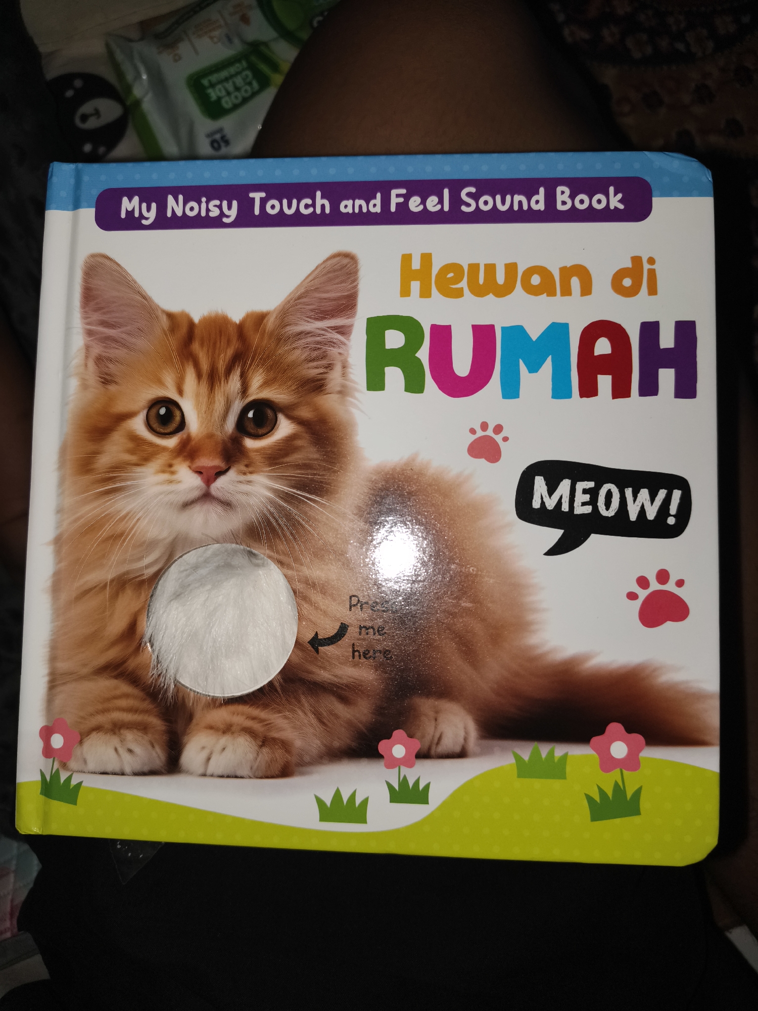 The image shows the cover of a children's book titled "My Noisy Touch and Feel Sound Book: Hewan di RUMAH". The cover features a fluffy orange kitten with large, expressive eyes. To the right of the kitten, a speech bubble contains the word "MEOW!". Below the kitten, there is a circular cutout with white fluffy material, labeled with an arrow pointing to it and the text "Press me here". The background is white, with decorative elements of pink paw prints, pink flowers, and green grass. The book is displayed on a dark surface, possibly a table or lap, and the background behind the book appears to be a blurry fabric. The overall impression is a colorful and engaging book for young children. The image does not provide any information about the city of Barengkrajan, Indonesia, or any specific location within it.