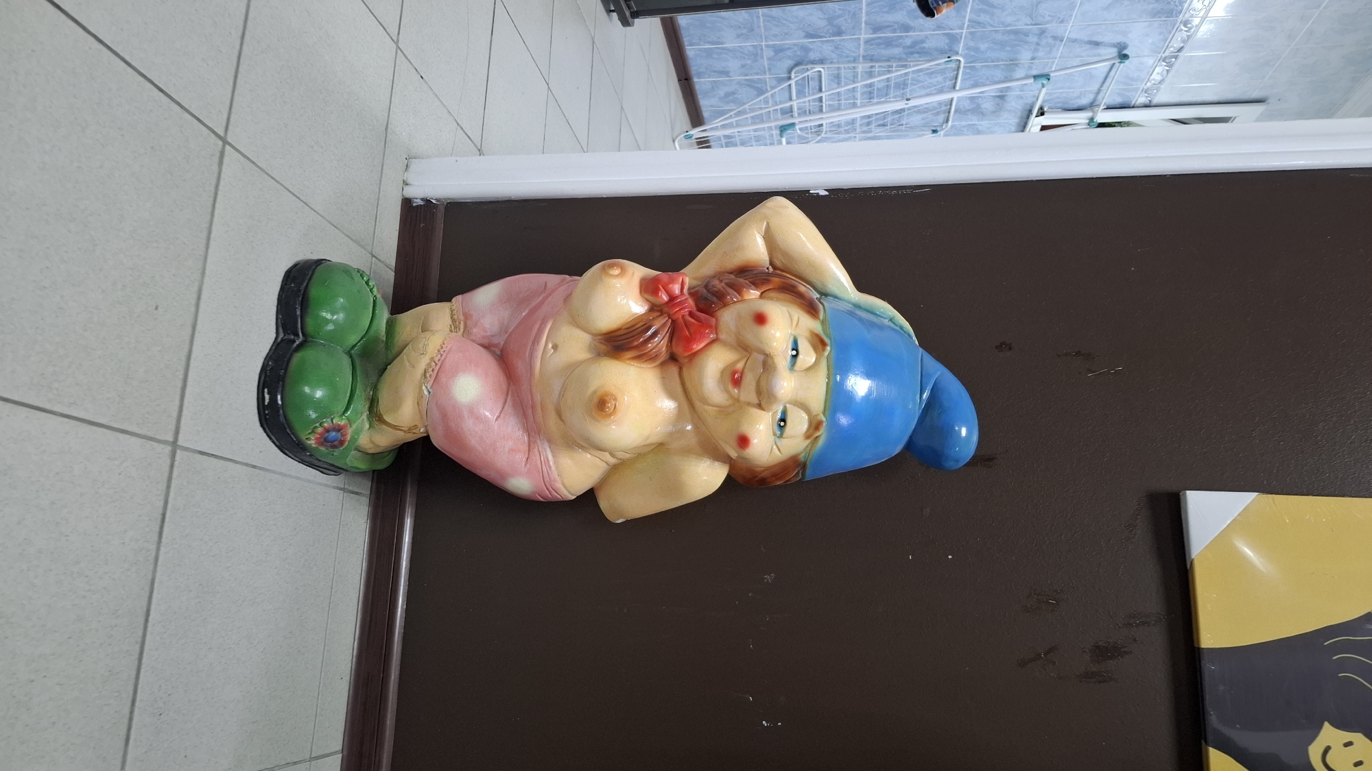 A statue of a scantily clad gnome stands in what appears to be an interior space. The gnome is depicted with a blue hat, rosy cheeks, and is wearing pink polka-dotted shorts and green shoes. It is positioned next to a dark brown wall. To the left of the gnome, a tiled wall with blue square tiles is visible, along with a white drying rack. The floor is covered with light-colored square tiles. The image does not provide any specific information or context related to the city of Niukzha, Russia.
