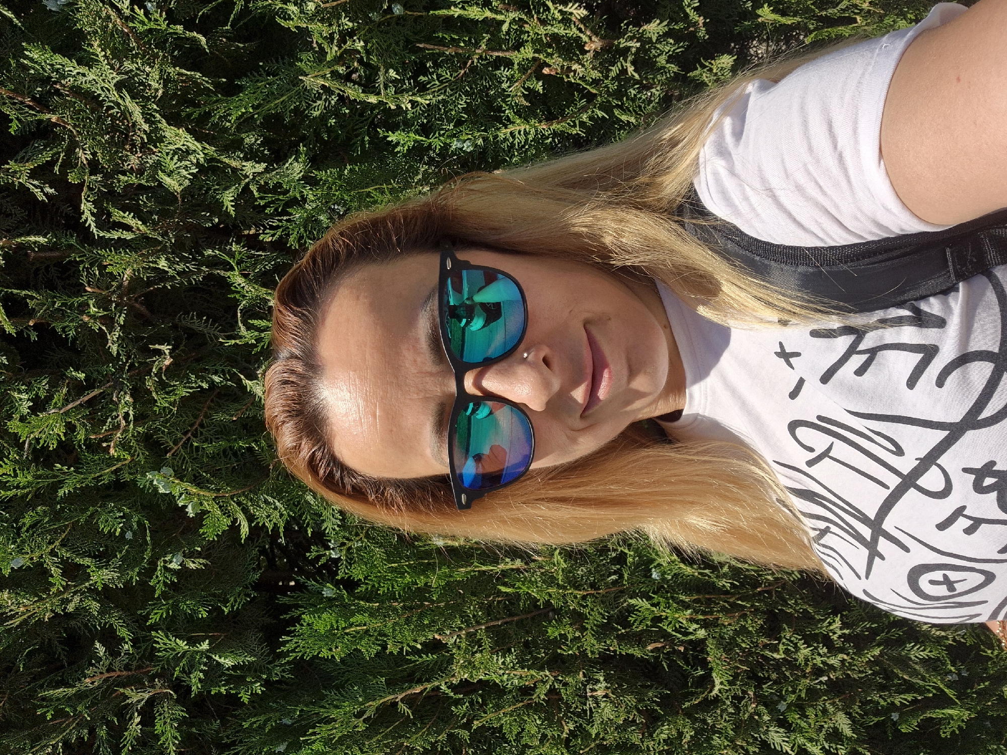 A woman with blonde hair and sunglasses is taking a selfie in front of a green hedge. She is wearing a white t-shirt with black graffiti-style writing on it and a black backpack. The woman is smiling slightly. The lighting suggests it is daytime.