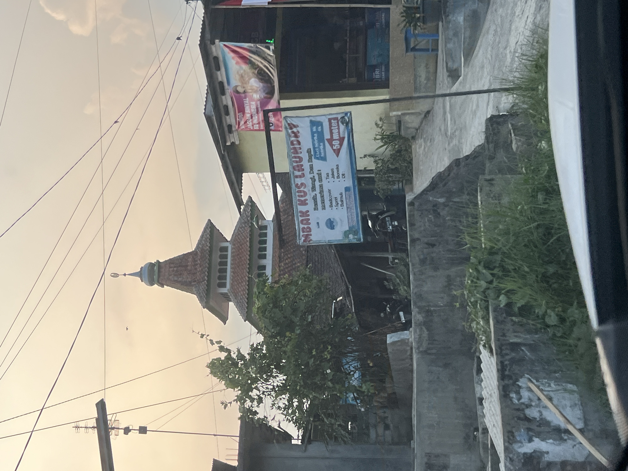 The image captures a street scene in Kapulogo, Indonesia, bathed in the warm glow of what appears to be late afternoon or early morning light. Dominating the background is the distinct tiered roof and minaret of a mosque, with a small crescent moon symbol atop its spire. To the right of the mosque, a yellow building houses a laundry service, indicated by a prominent sign reading "MBAK KUS LAUNDRY" which also lists services such as "Badcover, Sprei, Selimut, Tas, Jaket, Boneka, and Dill." A banner with an image of a woman and text in Indonesian, "JAGUAN SINYAL SE-INDONESIA," is also displayed on this building. A tree with dense green foliage stands to the left of the mosque. In the foreground, a concrete barrier and overgrown grass line the street, suggesting a somewhat unkempt but lived-in urban environment. Electrical wires are strung across the sky, and a bird is visible flying in the distance. The perspective suggests the photo was taken from a vehicle.