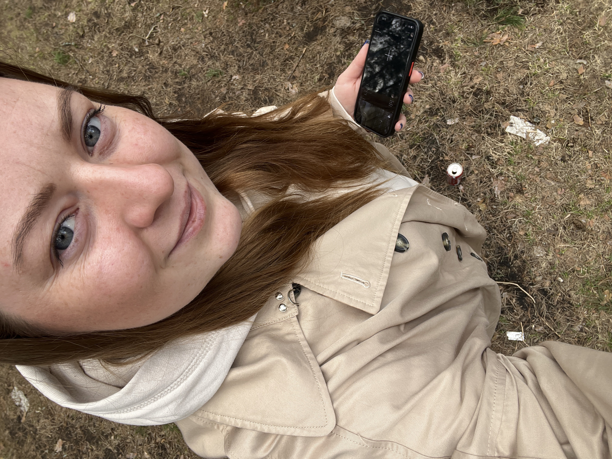 A woman with red-brown hair and light eyes is holding a black smartphone with a white cross on the screen in her right hand. She is wearing a beige trench coat over a white hooded sweatshirt. The photo appears to be a selfie, with the camera pointing down at her. The ground is covered with dry grass, leaves, and debris, including a discarded soda can and a piece of white trash. The location is Razdolie, Russia.