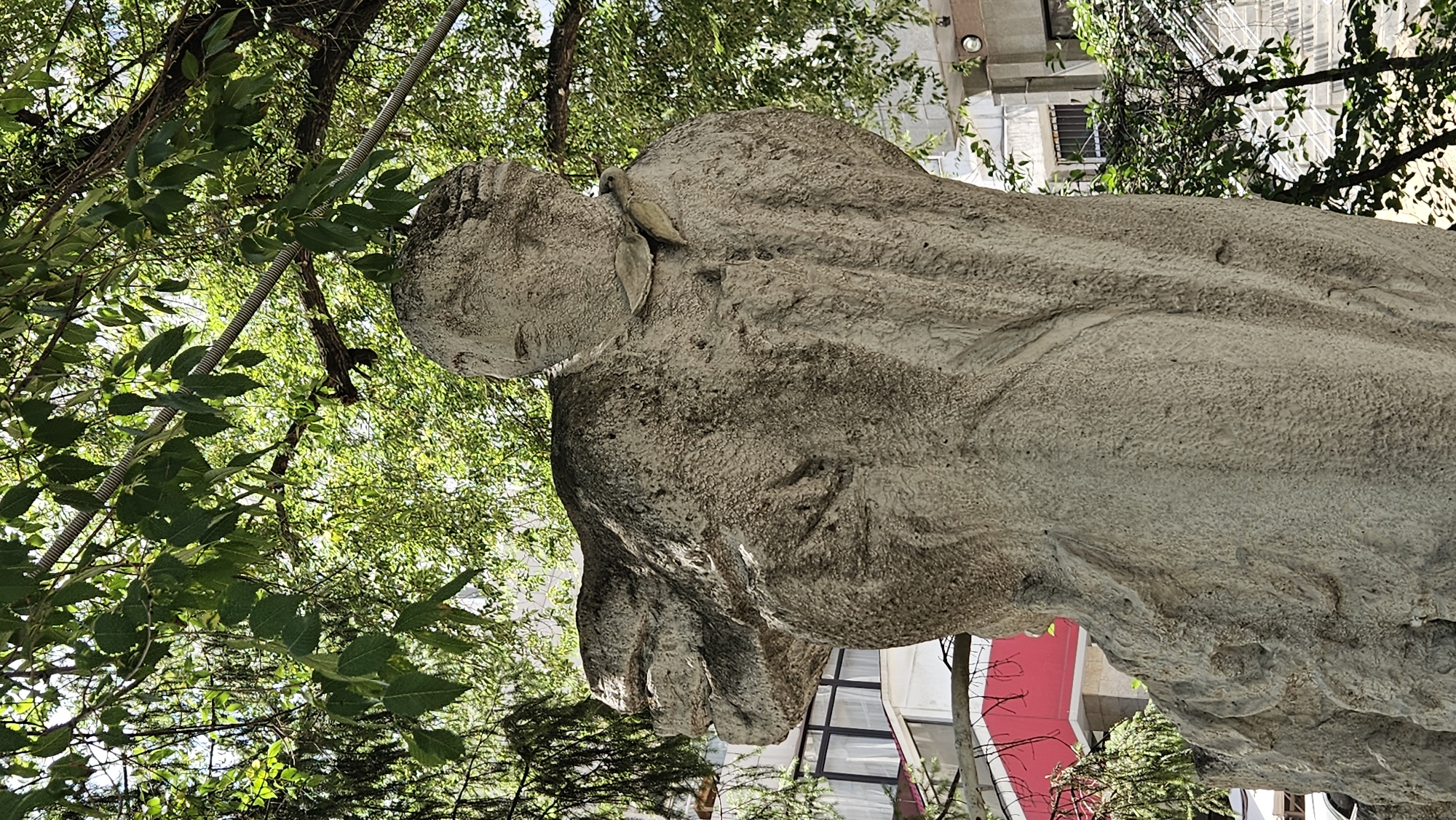This is a medium shot, low-angle, eye-level outdoor photograph of a rough, textured concrete sculpture. The sculpture depicts a human-like figure with a round head and outstretched arm. The figure is facing to the right, with its face turned slightly upwards. The surface of the sculpture has a coarse, gritty texture, and some areas appear darker, suggesting weathering or moss growth.
The sculpture is situated in an urban environment, likely in Almaty, Kazakhstan, as indicated by the surrounding buildings and vegetation. Behind the sculpture, there is a multi-story building with white and grey facades, featuring windows with dark frames and metal bars. A red awning or structure is visible on a lower level of the building. Green trees with dense foliage partially obscure the background, creating dappled light and shadow on the sculpture. Some of the tree branches extend over the sculpture, and a metallic pipe or conduit runs horizontally through the branches. The ground beneath the sculpture is not visible. The overall impression is that of an outdoor public space, possibly a park or a plaza, where the sculpture serves as a focal point.
