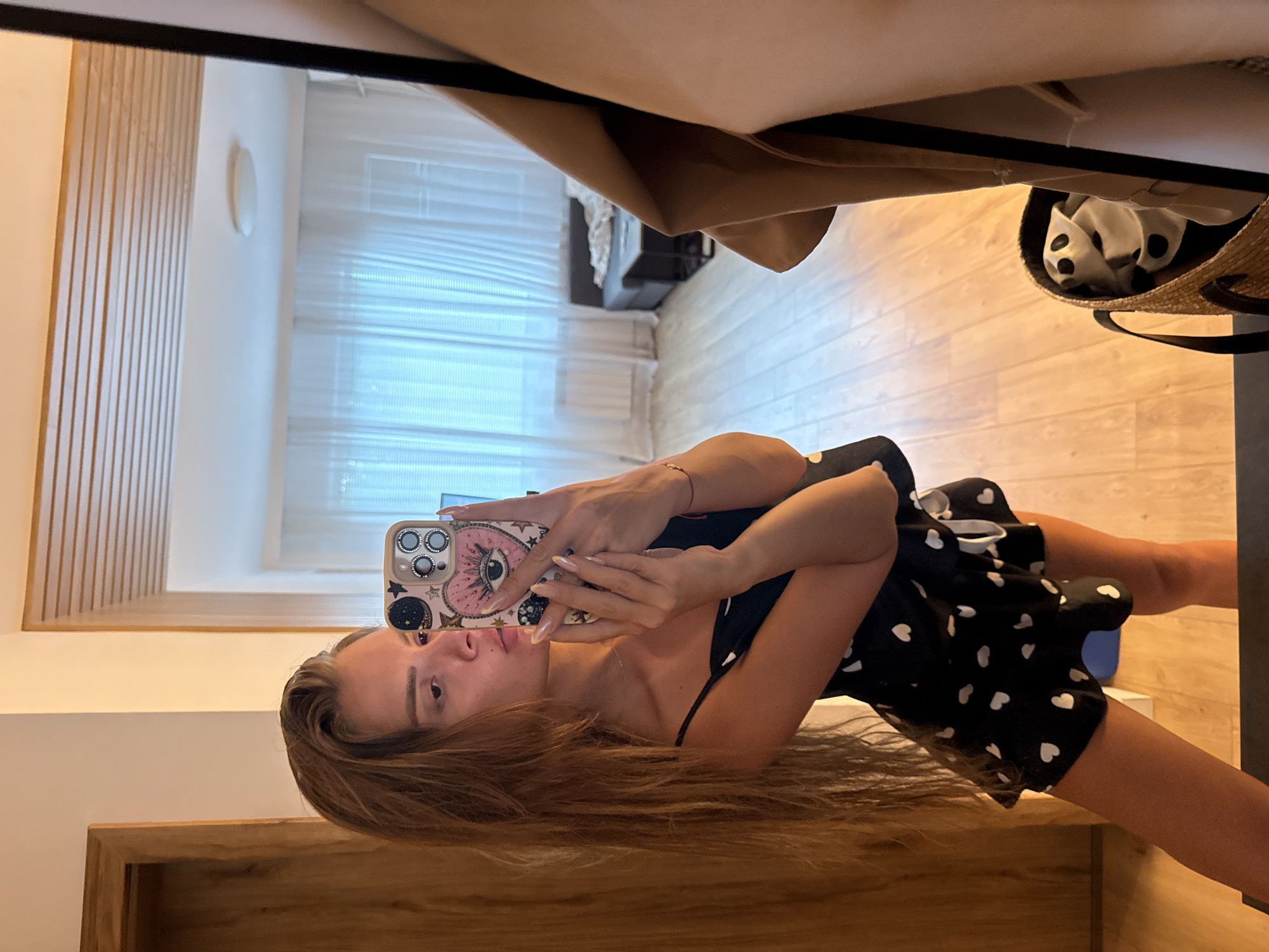 A young woman is taking a mirror selfie in what appears to be a hotel room or apartment. She is wearing a black pajama set with a white heart pattern. She is holding a phone with a heart-shaped case, which has an eye design on it. The room has wooden floors and white curtains in the background. A couch and a coffee table are visible in the reflection of the mirror. A straw bag with some items inside is on the floor in the foreground. The woman has long, brown hair and is wearing light makeup. Her fingernails are painted a shimmery pink. There is a jacket hanging on a rack to the right of the frame. The image does not contain enough information to determine a specific location in Plovdiv, Bulgaria.