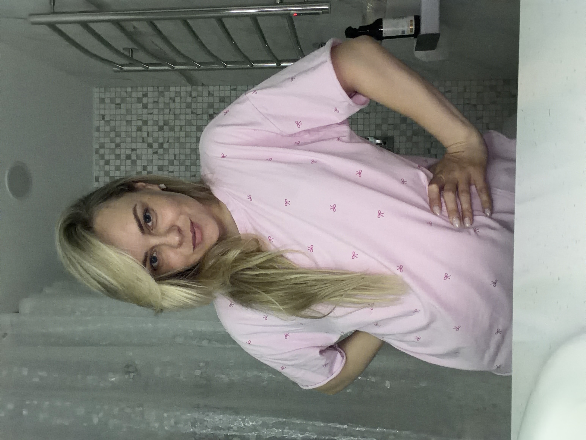 A woman with blonde hair is in a bathroom. She's wearing a pink t-shirt with small bow patterns. The bathroom has gray mosaic tiles and a silver towel rack. There's a shower curtain visible. The location in the city of Votkinsk, Russia is unknown.