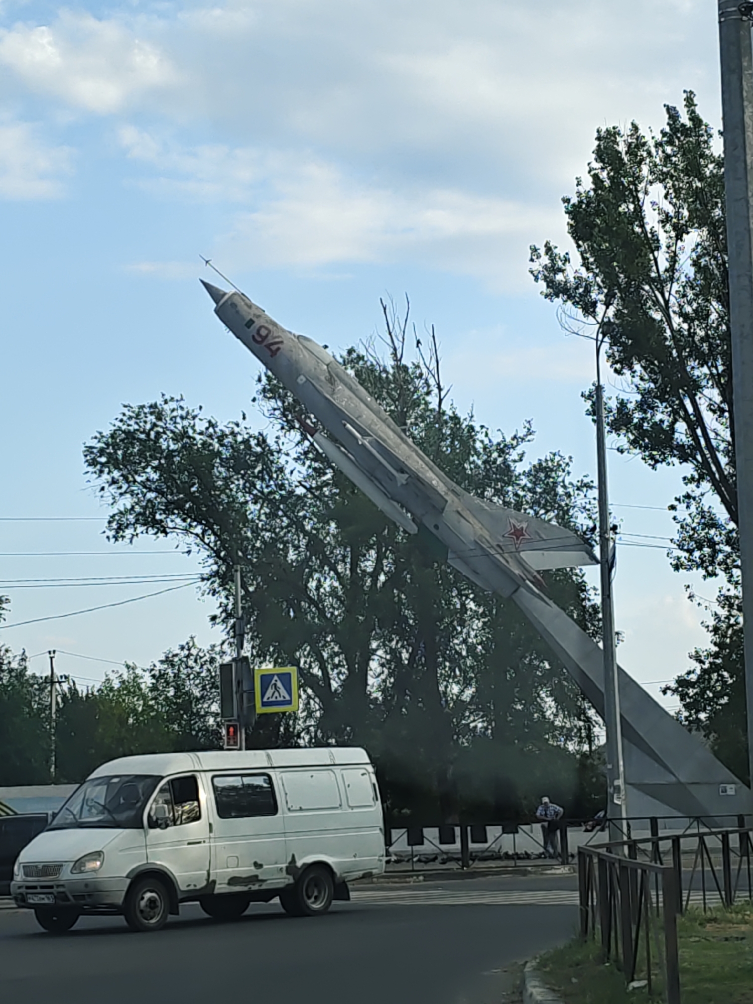 In Novocherkassk, Russia, a monument of a Soviet-era fighter jet is displayed outdoors, angled upwards against a sky with scattered clouds. The jet, painted gray, has the number "94" prominently visible on its fuselage. A white van is traveling on the road in front of the monument. Trees and utility poles are present in the background. A pedestrian crossing sign is visible near the jet’s base. One person is standing near a fence in the background.