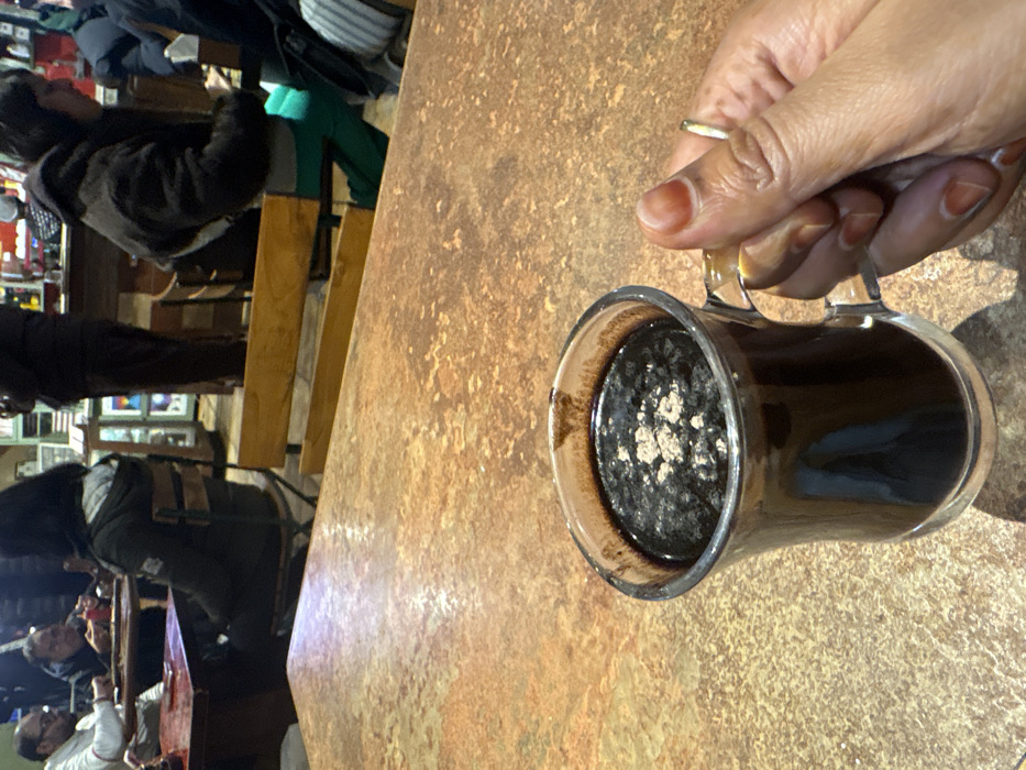 The image captures an indoor scene, likely in a cafe or restaurant, presented from a slightly overhead, diagonal perspective looking down onto a table. In the immediate foreground, a person's right hand is prominently featured, holding a clear glass mug. The hand has a warm, brownish skin tone, and a simple gold-colored ring is visible on the ring finger. The fingernails are short and appear to have a reddish hue, possibly from henna.
The glass mug contains a dark, rich-colored beverage, resembling hot chocolate or coffee, with a distinct layer of lighter-colored, frothy or powdered topping on its surface. This topping, which appears speckled and unevenly distributed, suggests additions like cocoa powder, cinnamon, or a milk foam. The mug itself has a handle, which the hand is gripping.
The mug and hand are resting on or hovering above a sturdy table with a textured, mottled surface. The table's color scheme consists of various shades of brown, tan, and rust, giving it a somewhat rustic or stone-like appearance.
In the blurred background, several other individuals are visible, indicating a social setting. They are seated at other tables or benches. One person in the mid-ground, slightly to the left, has dark hair and is wearing a dark, possibly fuzzy, jacket over what appears to be green pants or a garment with green stripes. Other figures are less distinct, but their presence suggests a lively, public environment. Wooden benches or seating structures are also discernible in the background. The overall lighting appears to be artificial, creating some reflections on the table and the mug. There is no visible text in the image.