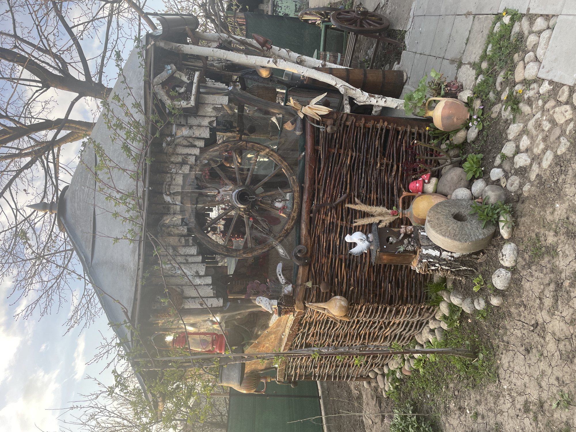 The image shows a cluttered outdoor scene in Aprilovo, Bulgaria. A small, shed-like structure with a sloped, gray roof is centrally located. The structure's facade incorporates various repurposed materials, including birch logs, a wooden wagon wheel, and a woven branch fence. Decorative items are arranged in front of the fence, including pottery jugs, a mushroom ornament, and a stone mill wheel. The ground surrounding the structure is a mix of cracked earth, gravel, and vegetation. In the background, a wooden wagon wheel and green fence are partially visible. Bare tree branches are visible at the top of the frame against a pale sky.