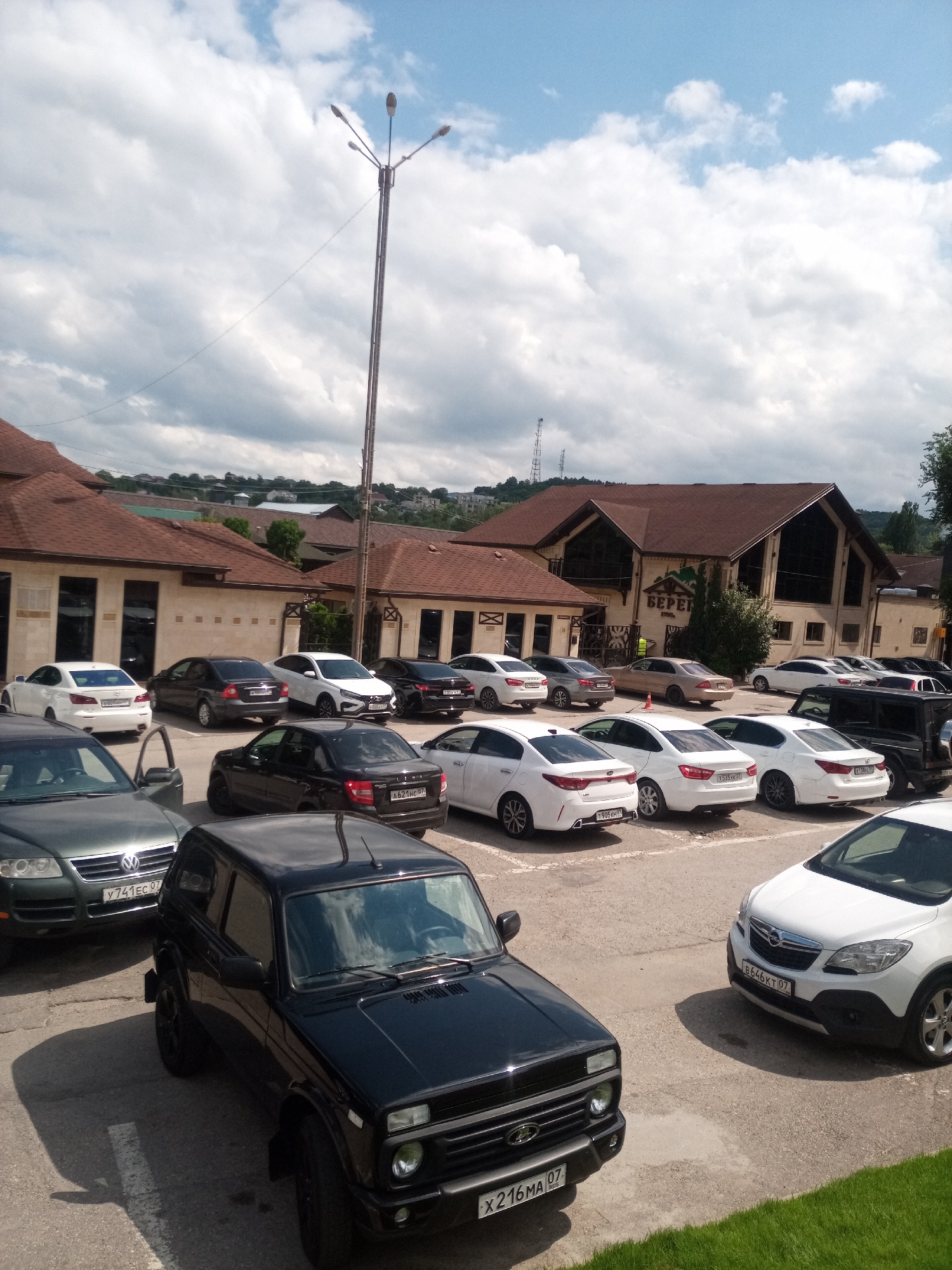 A daytime, high-angle, eye-level medium shot captures a parking lot filled with various vehicles and a building complex in Nalchik, Russia. The sky above is a mix of blue with scattered clouds.
In the foreground, a black Lada Niva is parked, its front passenger door open. To its left, a dark green Volkswagen Touareg is visible, with a license plate reading "Y 741 EC 07". Further to the right and slightly behind the Lada, a white Opel Mokka is parked, displaying a license plate "B646KT 07".
In the mid-ground, a variety of cars are parked in rows. These include white Kia Rio, white Hyundai Solaris, and several dark-colored sedans. A black Mercedes-Benz G-Class is also visible in the mid-ground.
The background features a multi-building complex with brown-tiled roofs and beige-colored walls. One building has a sign with the Russian word "БЕРЕГ" (Bereh). A street light with multiple bulbs stands tall on the left side of the frame. The surrounding area appears to be a suburban or semi-rural setting, with trees and a gentle slope of buildings in the distance. The overall scene depicts a typical parking area at a commercial establishment on a clear day.