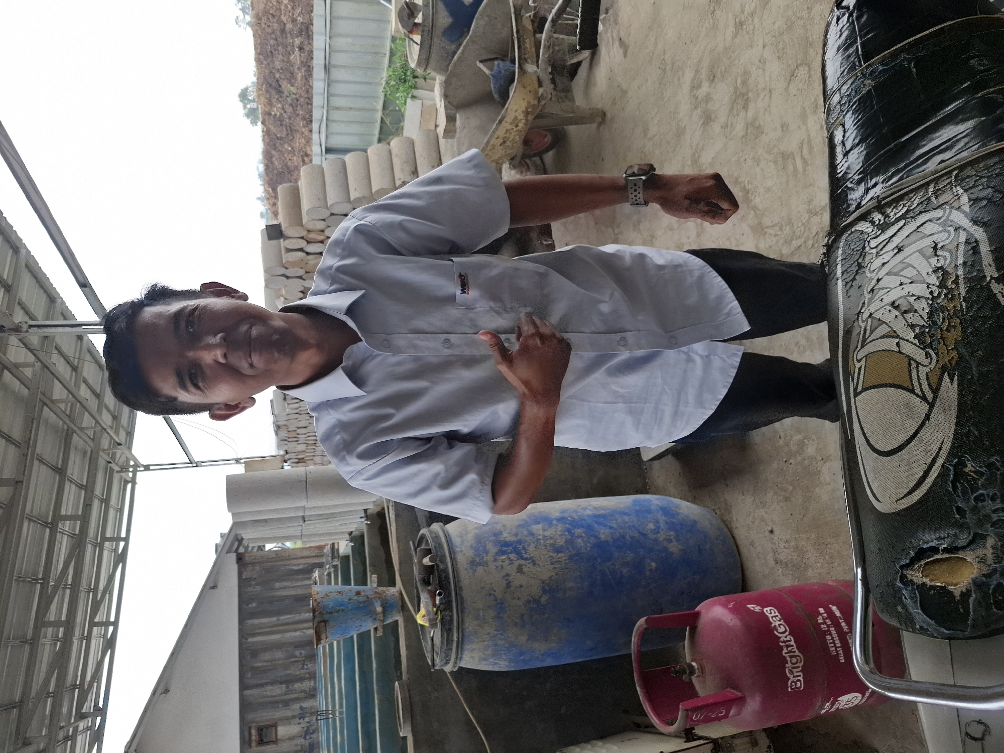 A man in a light blue collared shirt and dark pants stands in an outdoor industrial area, possibly a construction or manufacturing site, in Kutasirna, Indonesia. He is looking at the camera and gesturing with his left hand, which is partially visible. Behind him, to his left, is a large blue plastic barrel. To his right, there are stacks of cylindrical concrete or stone objects and a wheelbarrow. In the foreground, a red gas cylinder labeled "Bright Gas" is visible, and a portion of a damaged motorcycle seat is in the lower part of the frame. The background shows a building with a metal roof and a dirt embankment. The sky is overcast.