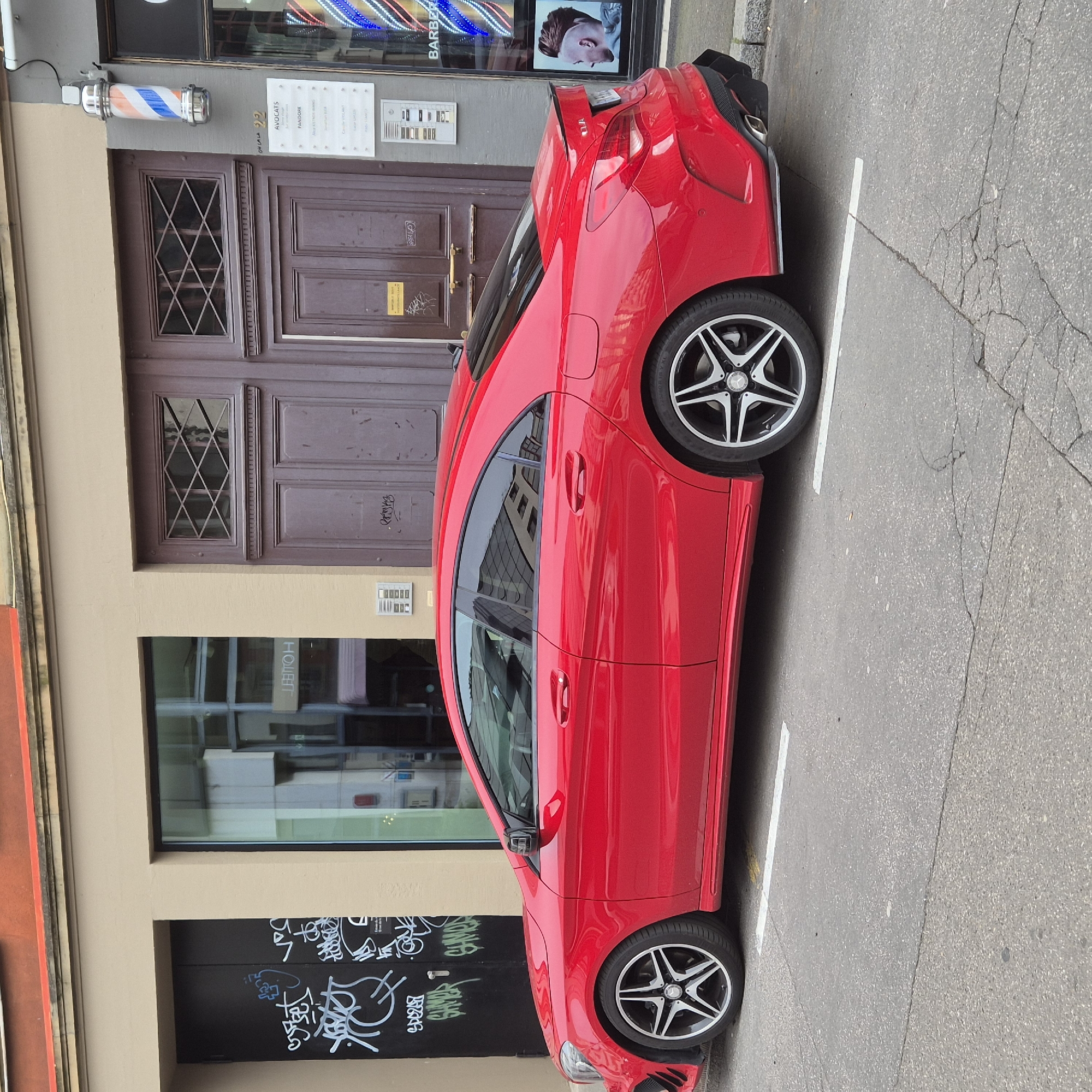 The image shows a red Mercedes-Benz CLA parked on a street in Strasbourg, France. The car is parked beside a building with a barber shop on the upper floors. The building has a wooden door with decorative glass and a black door with graffiti on the lower floors. The street is paved with asphalt. The car is parked between two white lines, indicating a designated parking space.