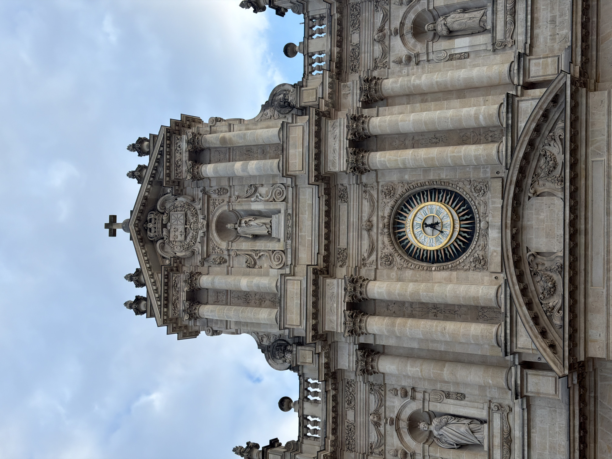 The image depicts the facade of a building, likely a cathedral, in Paris, France. The structure is made of light-colored stone. It features intricate architectural details, including columns, sculptures, and a clock. At the top is a cross. The sky is visible behind the building.
