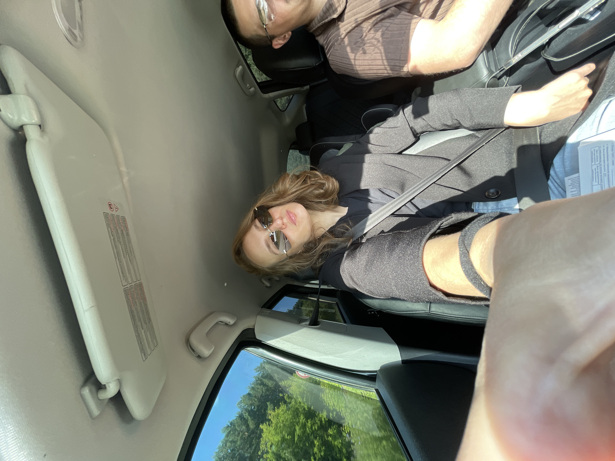 A woman wearing sunglasses and a black jacket is seated in the passenger seat of a car, holding up her left arm, likely to take a selfie. She is wearing a seatbelt. Next to her, a man wearing glasses and a brown shirt is in the driver's seat. The car's interior is visible, including the sun visor. Through the window, a treed landscape with a bright blue sky can be seen. The setting is likely within or near the city of Shud'ya, Russia, given the context of the image's origin.