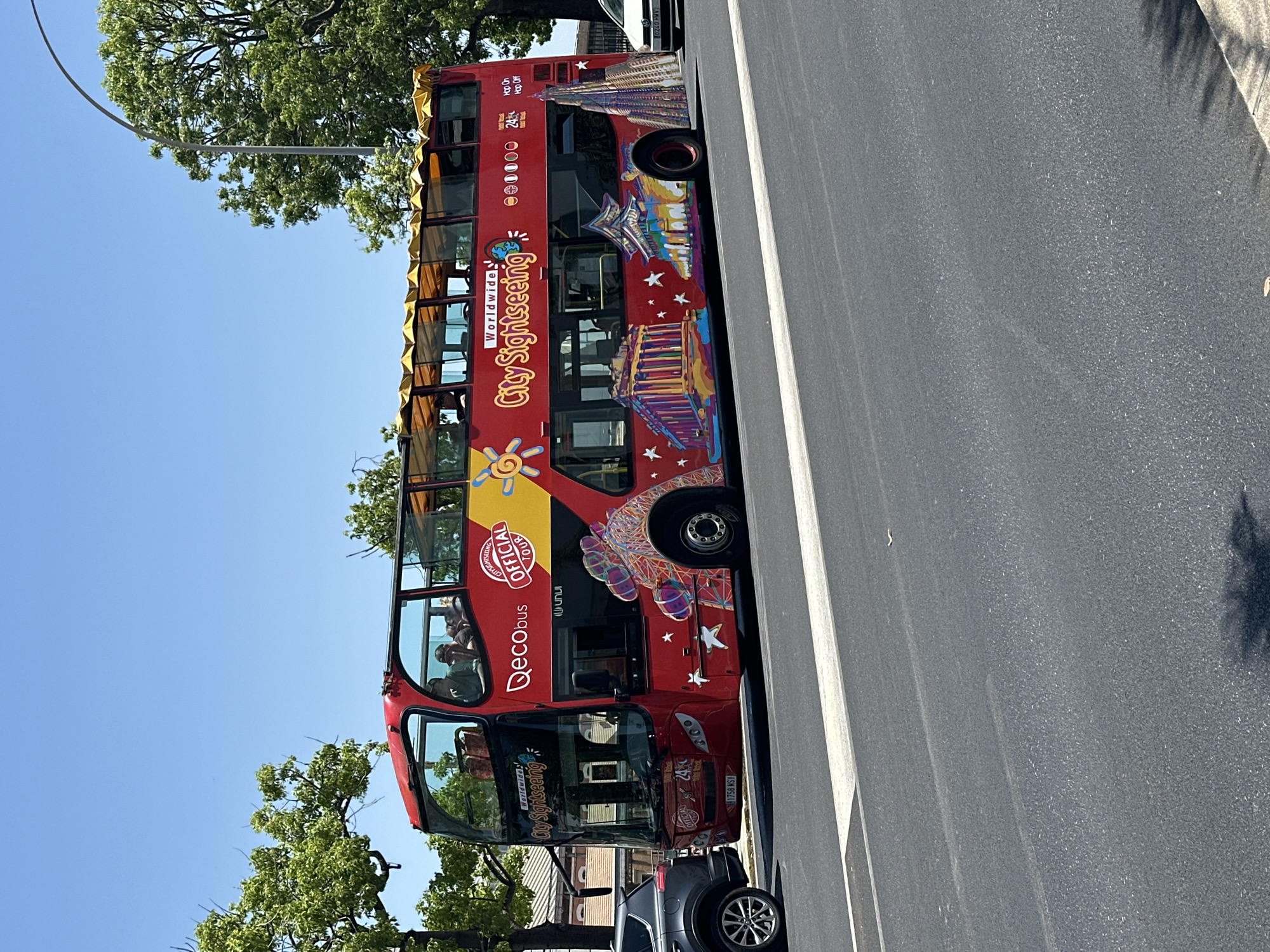 A red, open-top double-decker tour bus with the branding "Qecobus City Sightseeing Official Tour Worldwide" is parked on a city street. The bus is decorated with colorful images of various landmarks, including what appears to be the Parthenon, a pagoda, and a skyscraper. There are several passengers seated on the upper deck of the bus, enjoying the ride. In the background, trees and buildings suggest an urban environment. The street is paved, and a white line runs across it, indicating a pedestrian crossing or lane marking. To the left of the bus, a dark-colored SUV is visible. The sky is clear and blue, suggesting a sunny day. The license plate of the bus is partially visible as "1758 WSV". The image appears to have been taken in Cádiz, Spain, given the architectural styles depicted on the bus and the overall ambiance.